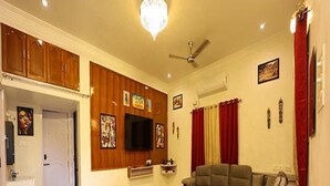 A living room with a sectional sofa, ottoman, wooden cabinets, a flat-screen TV, and artwork on the walls. - Fabescape Queens Paradise (Vanur)