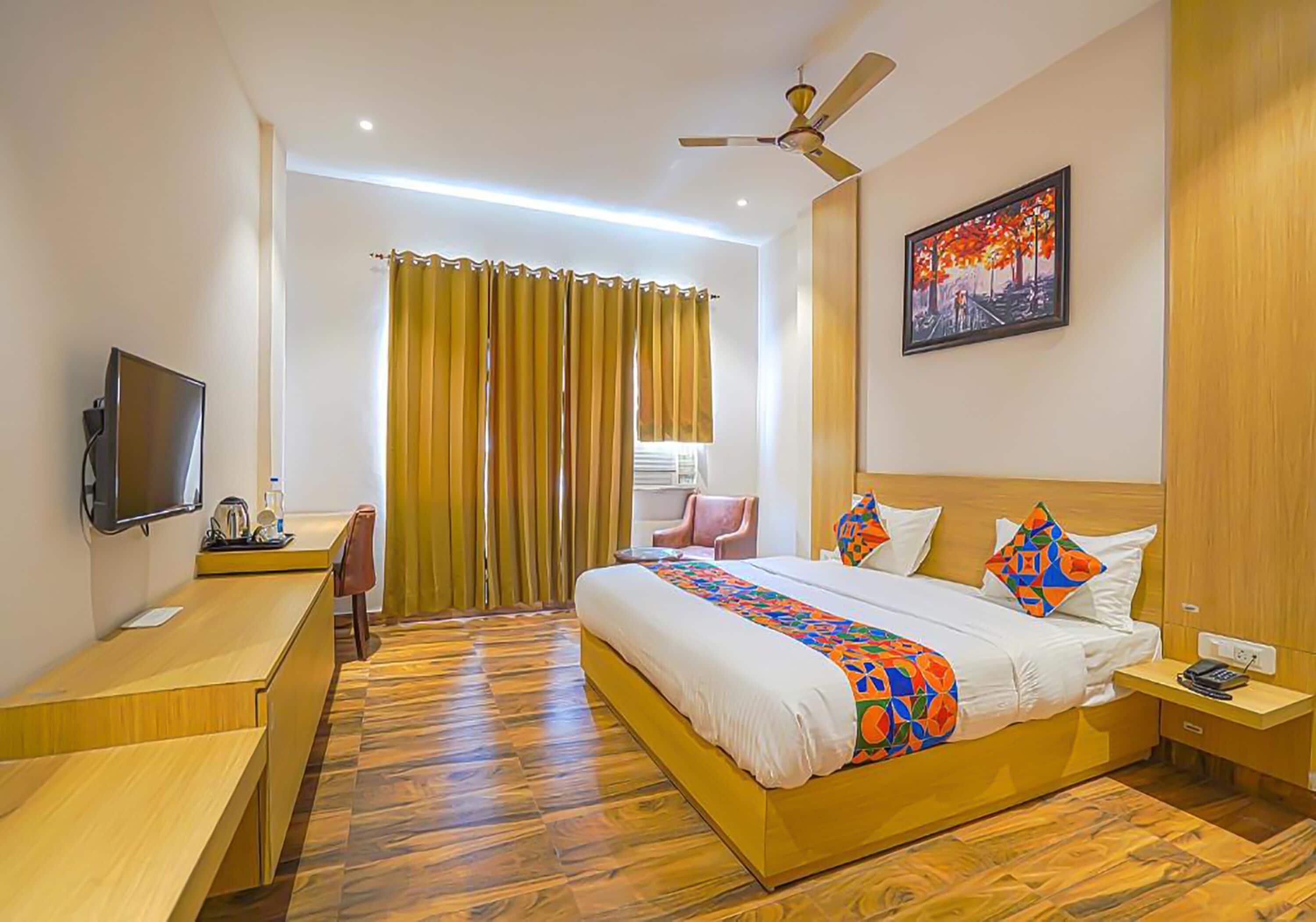 Une chambre d’hôtel avec un lit, un bureau, une télévision, un ventilateur de plafond et un tableau au mur.