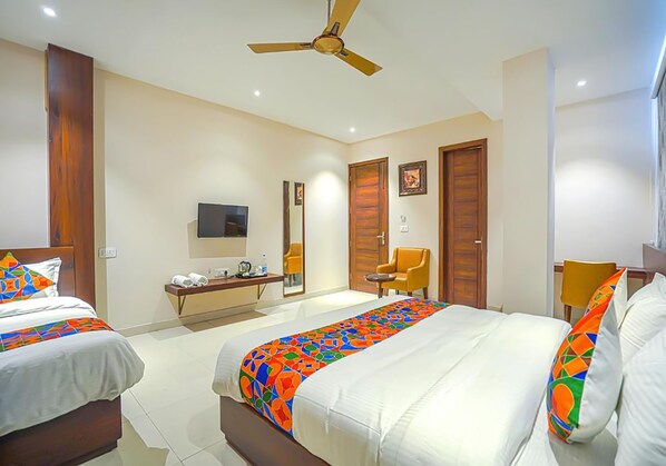 Habitación de hotel con dos camas, ventilador de techo, televisión, escritorio con silla, y baño con espejo e inodoro.