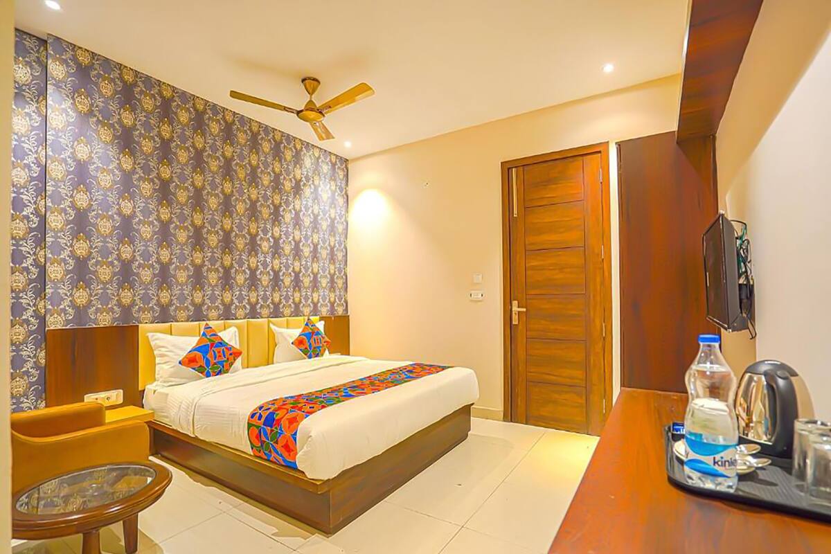 A hotel room with a bed, a desk with a water bottle and a kettle, a chair, and a ceiling fan.