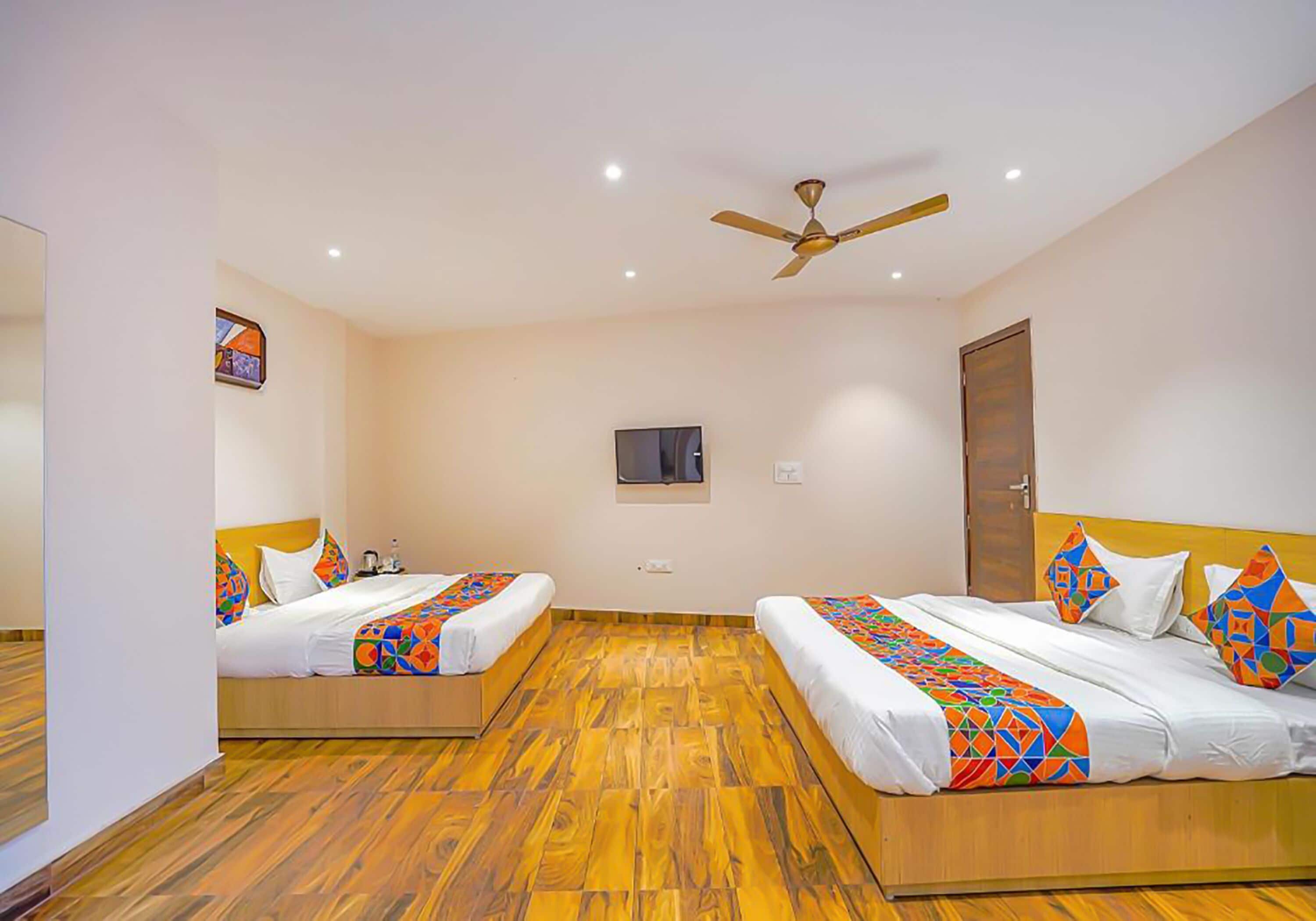 A hotel room with two beds, wooden flooring, a ceiling fan, and a television on the wall.
