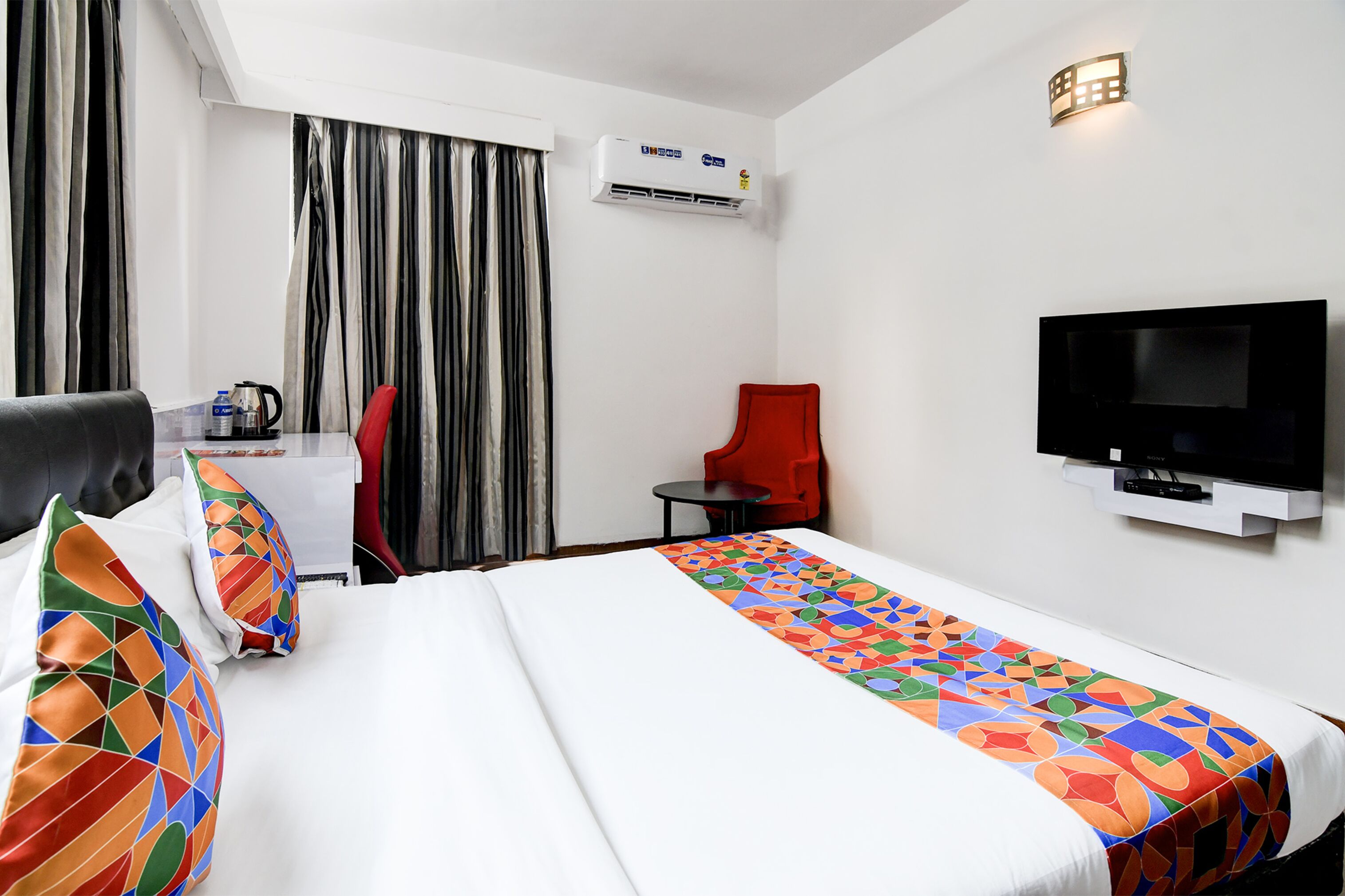 Habitación de hotel con cama, televisión, silla, mesita y aire acondicionado.