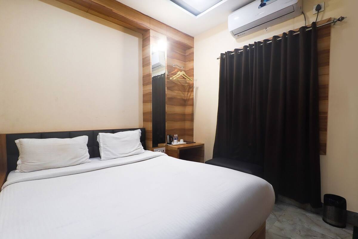 A hotel room with a bed, a nightstand, a wall-mounted air conditioner, and a window with curtains.