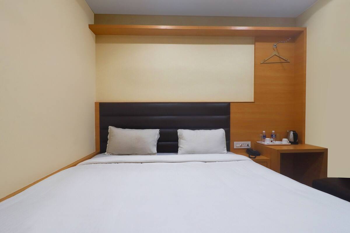 A hotel room with a bed, a wooden headboard, a bedside table with a phone and a kettle, and a hanging coat rack.