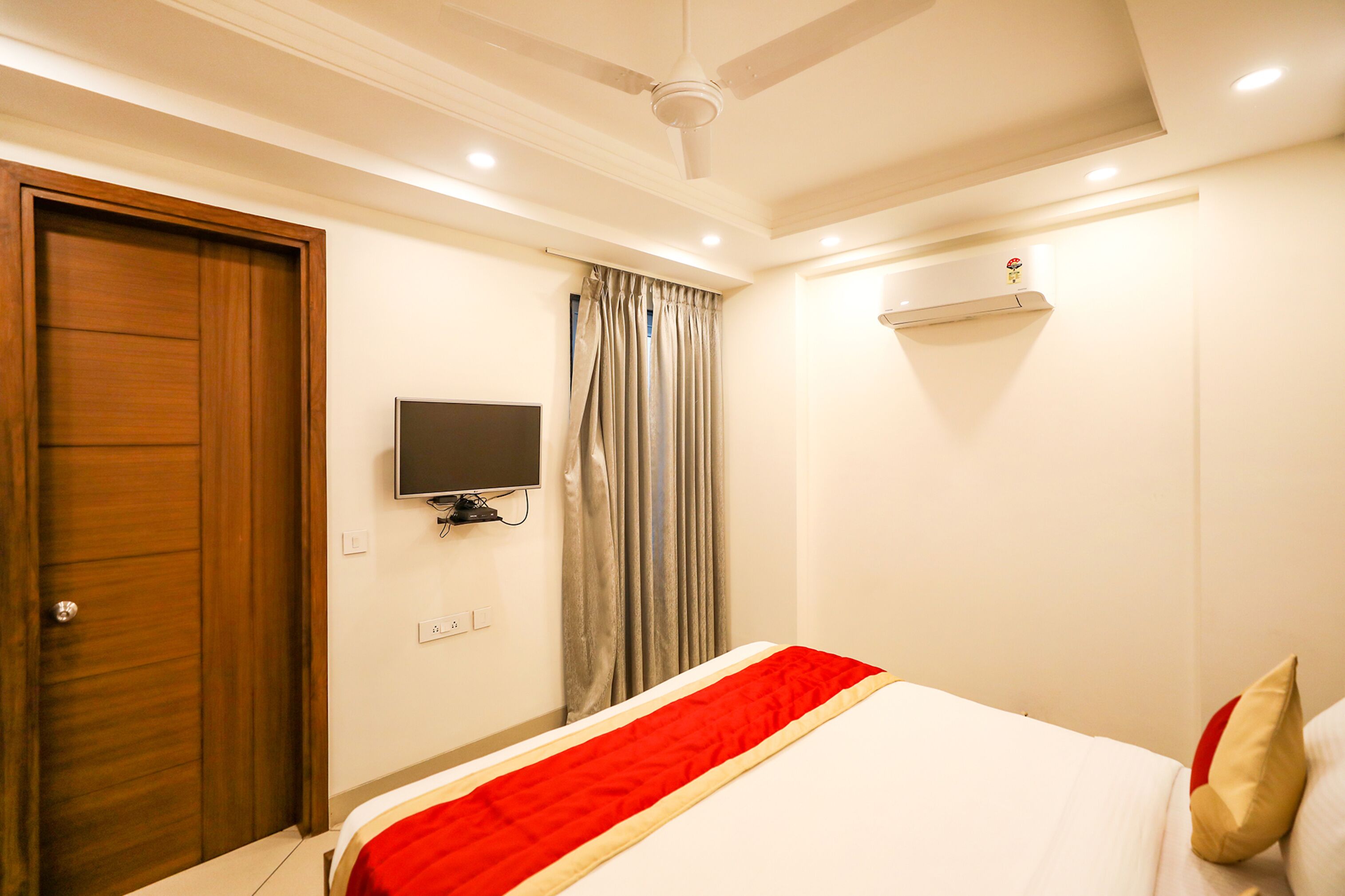 A hotel room with a bed, a television mounted on the wall, a wooden door, and a ceiling fan.