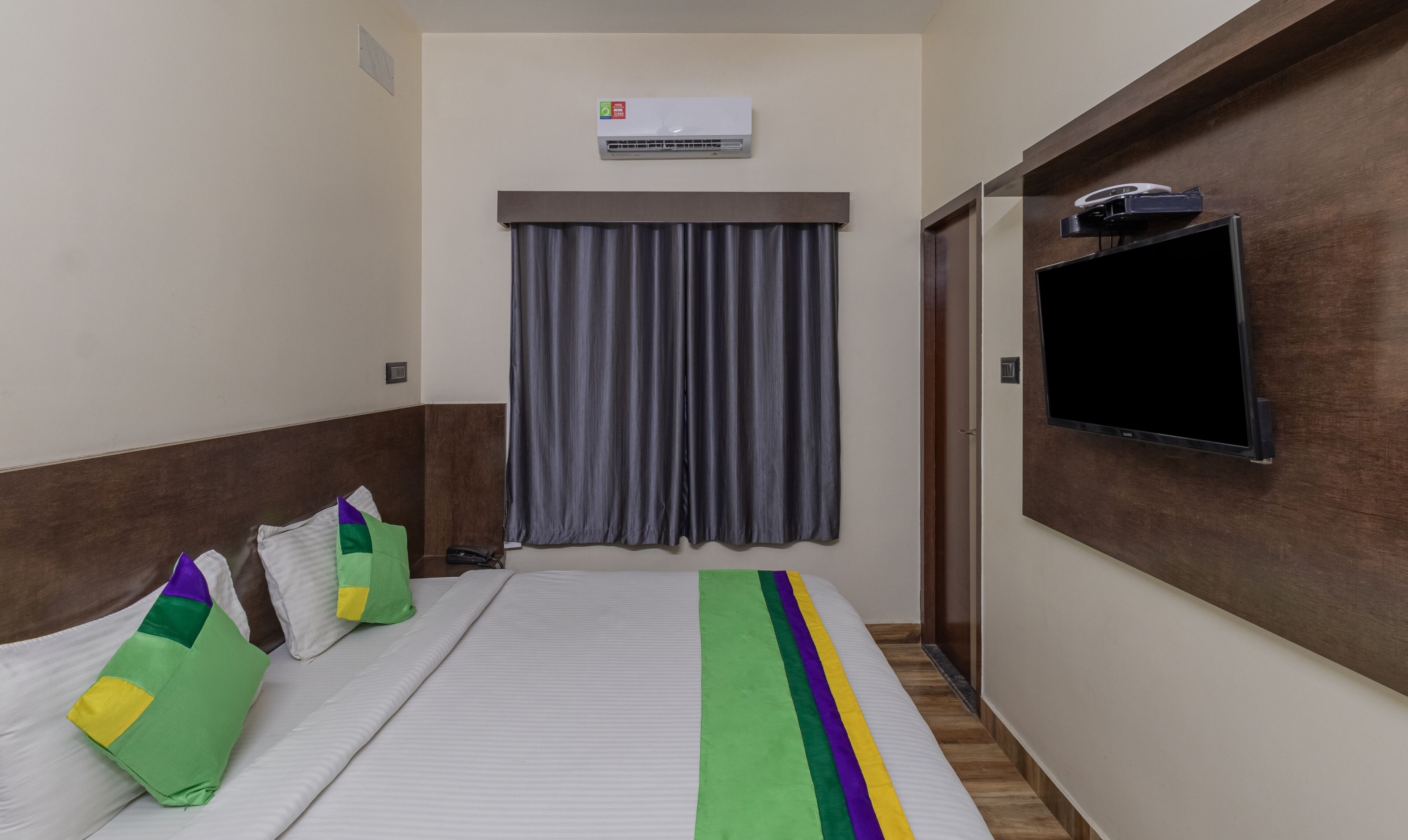 A hotel room with a bed, a TV mounted on the wall, an air conditioning unit, and a window with curtains.