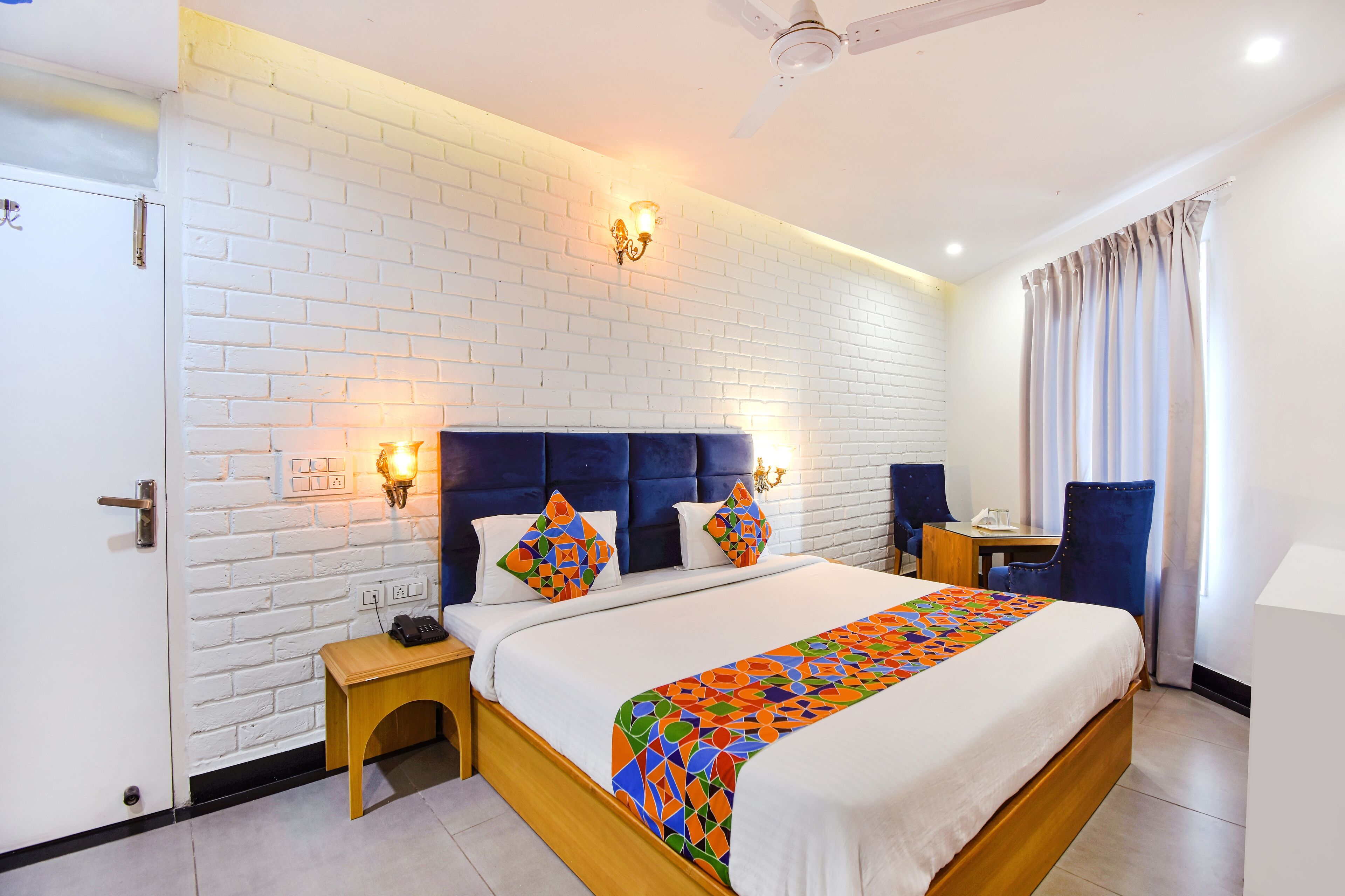 A hotel room with a white brick wall, a wooden bed with a colorful blanket, two bedside tables, and a desk with a chair.