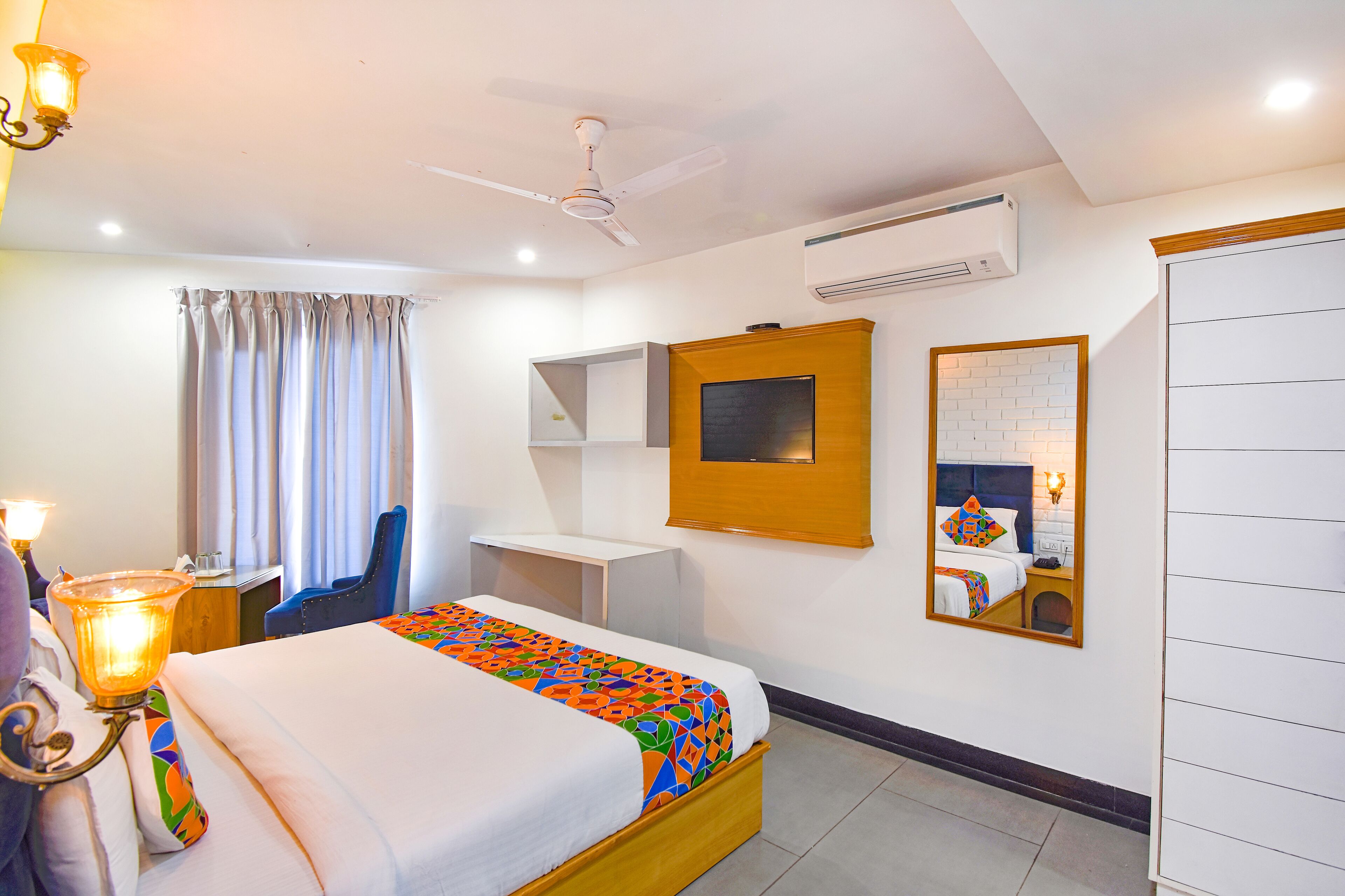 A hotel room with a bed, a desk, a chair, a ceiling fan, an air conditioning unit, and a mirror.
