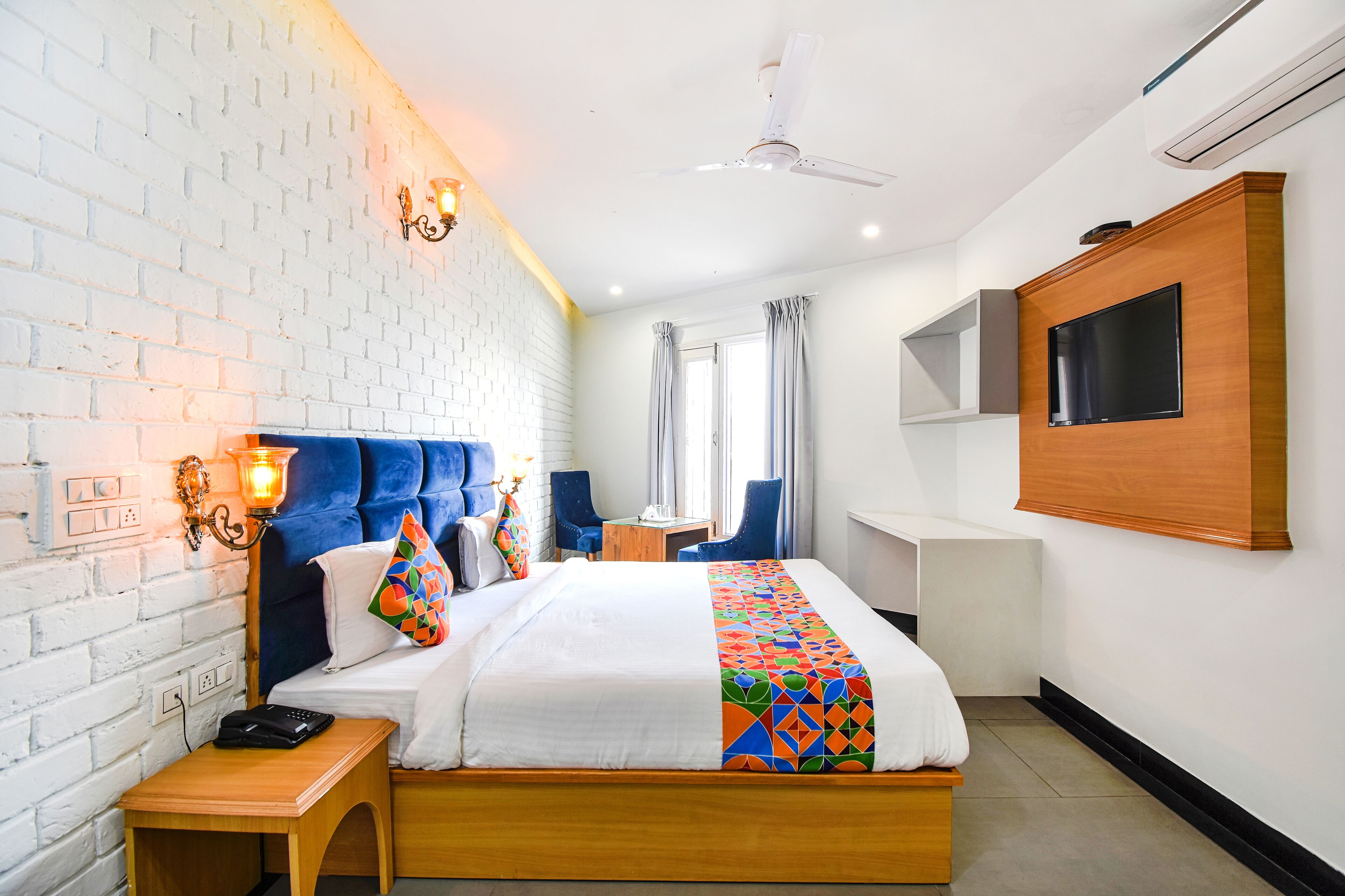 A hotel room with a brick wall, a bed with a colorful geometric bedspread, a desk with a chair, a TV mounted on the wall, and a ceiling fan.