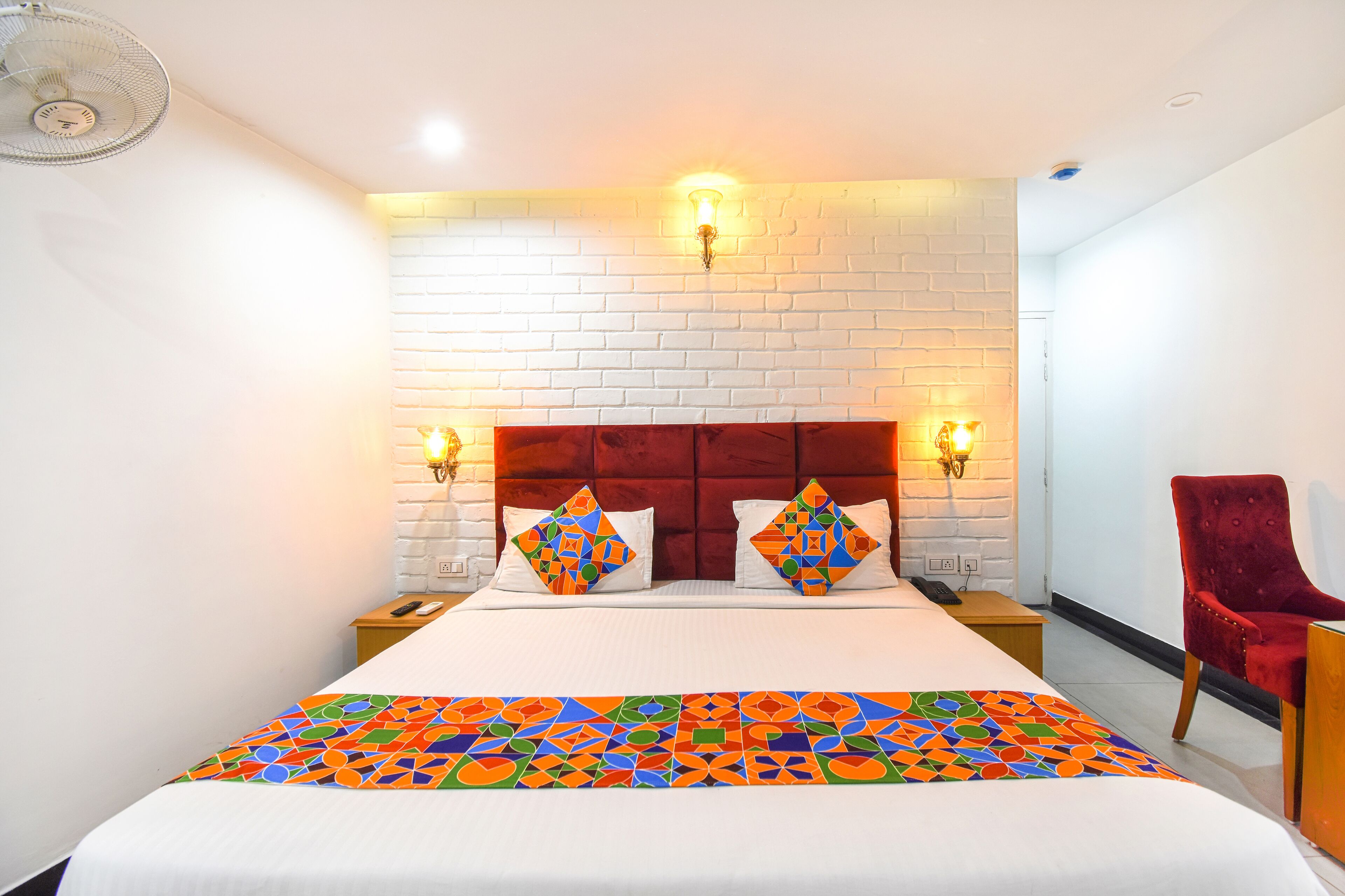 A hotel room with a white brick wall, a red upholstered headboard, two bedside lamps, a red chair, and a colorful bedspread.