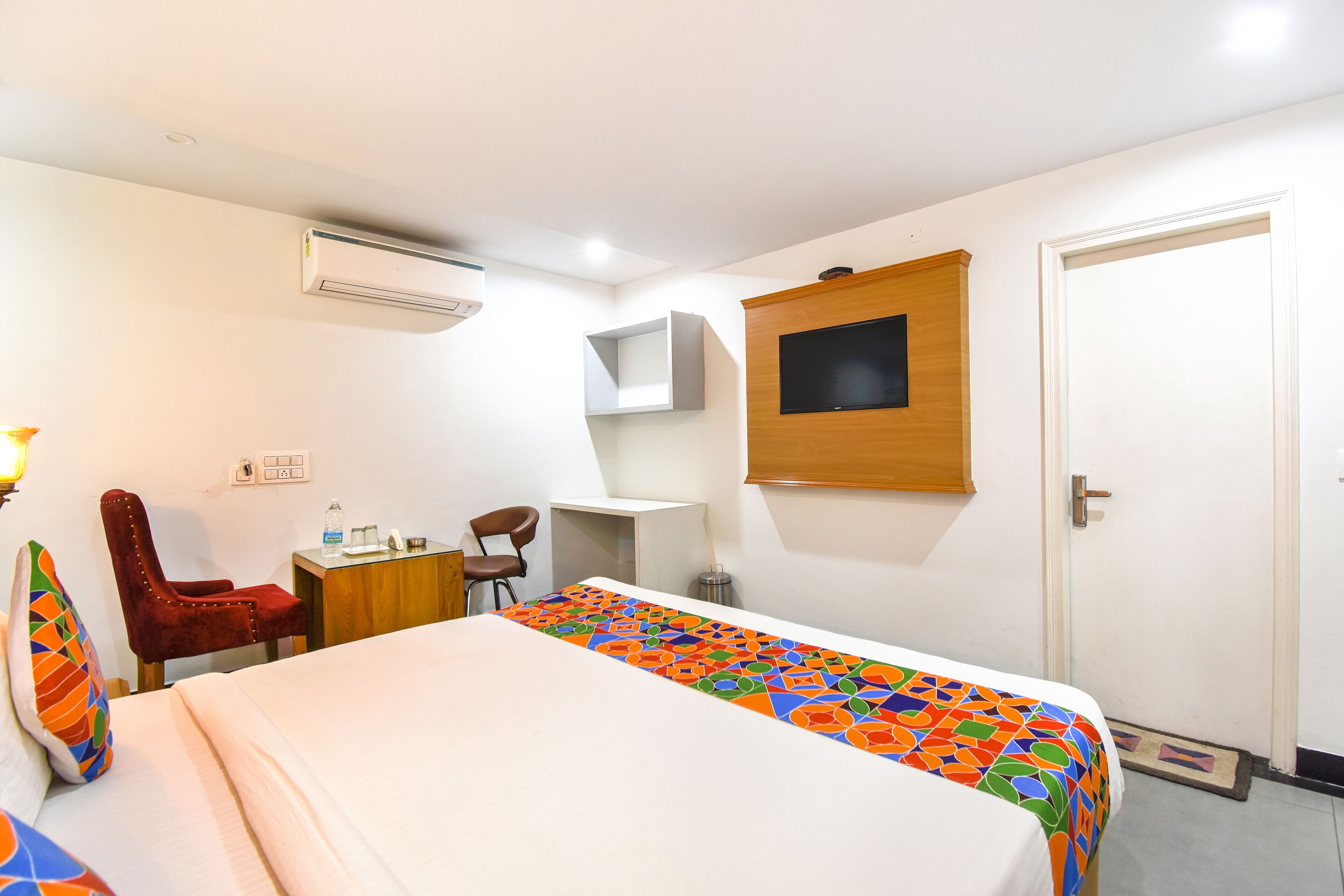 A hotel room with a bed, a desk, a chair, a TV, and an air conditioning unit.