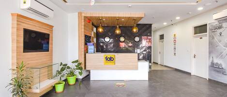 A hotel reception area with a wooden reception desk, air conditioning units, potted plants, and a corridor with doors.