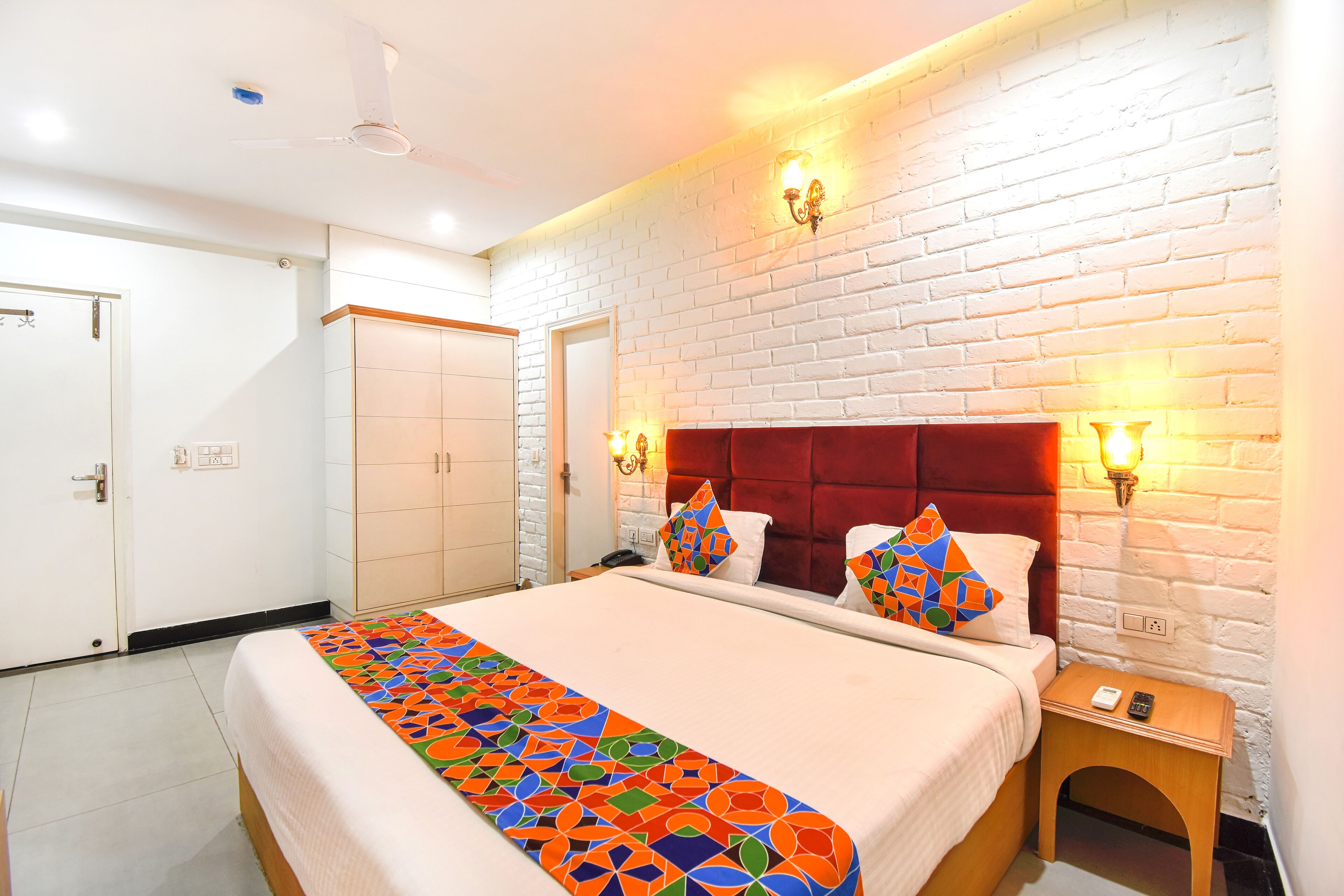 A hotel room with a large bed, a red headboard, white brick walls, and two bedside lamps.