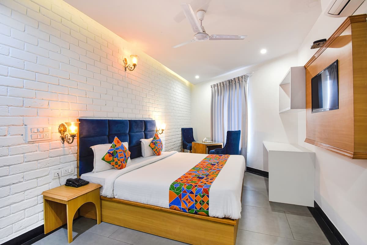 Een hotelkamer met een witte bakstenen muur, een houten bedframe, een blauw gestoffeerd hoofdeinde, een witte nachtkastje, een klein houten nachtkastje, een kleurrijke plaid, een flatscreen televisie aan de muur, een plafondventilator en een raam met gordijnen.