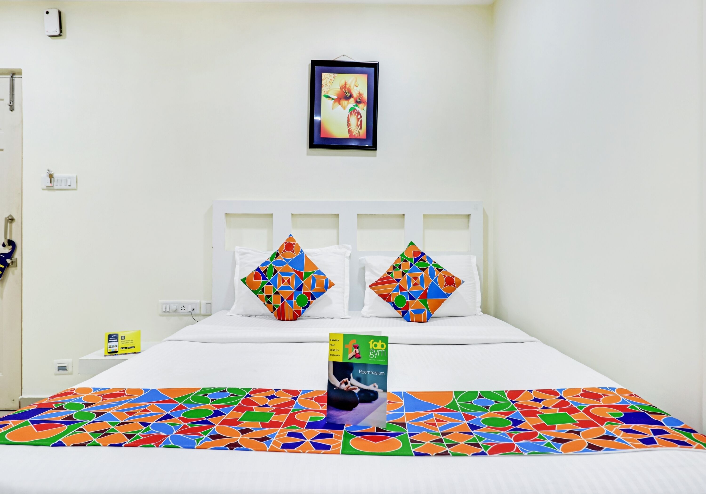 Una habitación de hotel con una cama, una colcha de diseño geométrico y colores vivos, dos almohadas con un patrón similar, un cuadro en la pared y una mesita con un letrero.