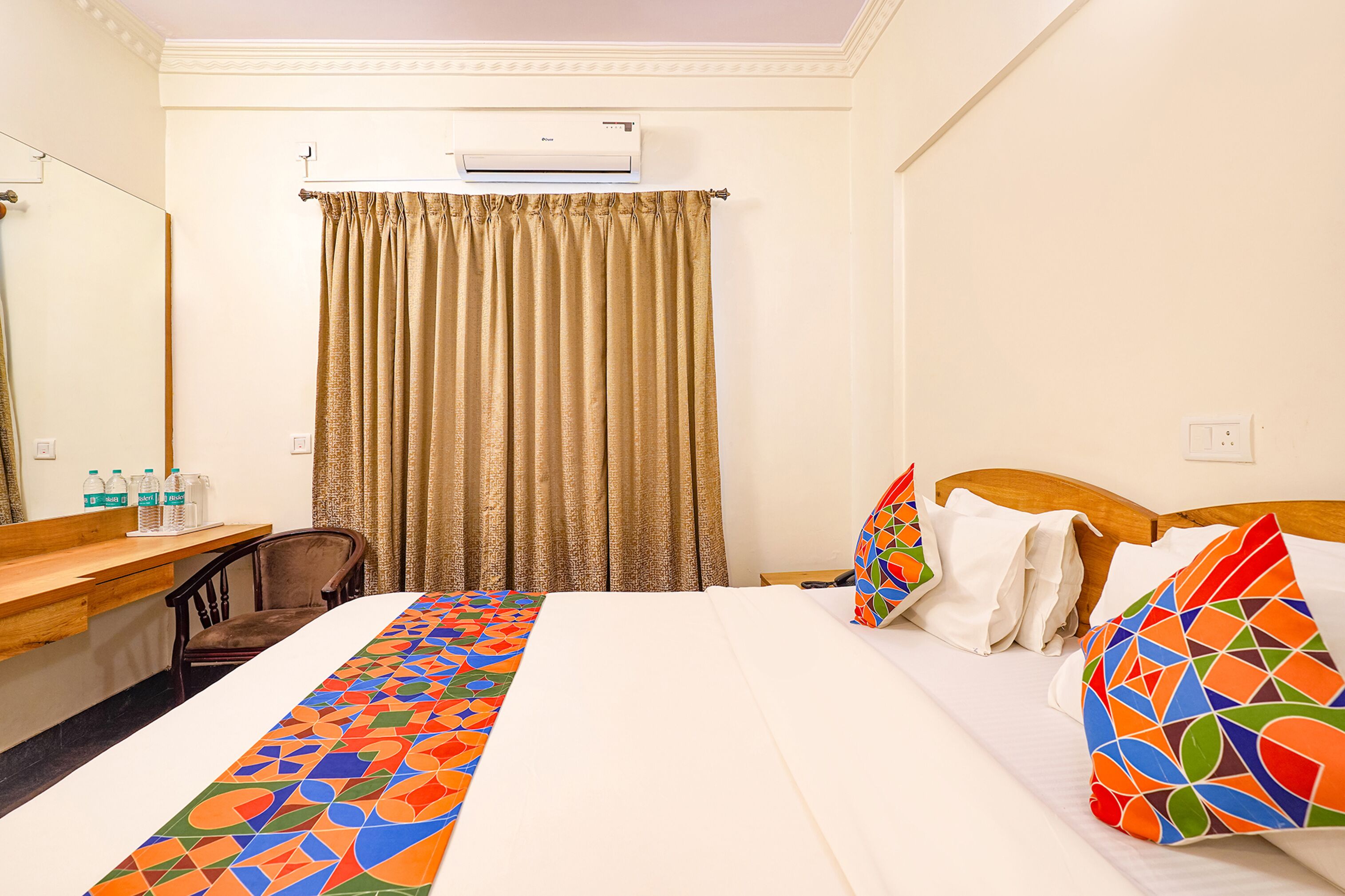 A hotel room with a bed, a desk with a chair, a mirror, and an air conditioning unit.