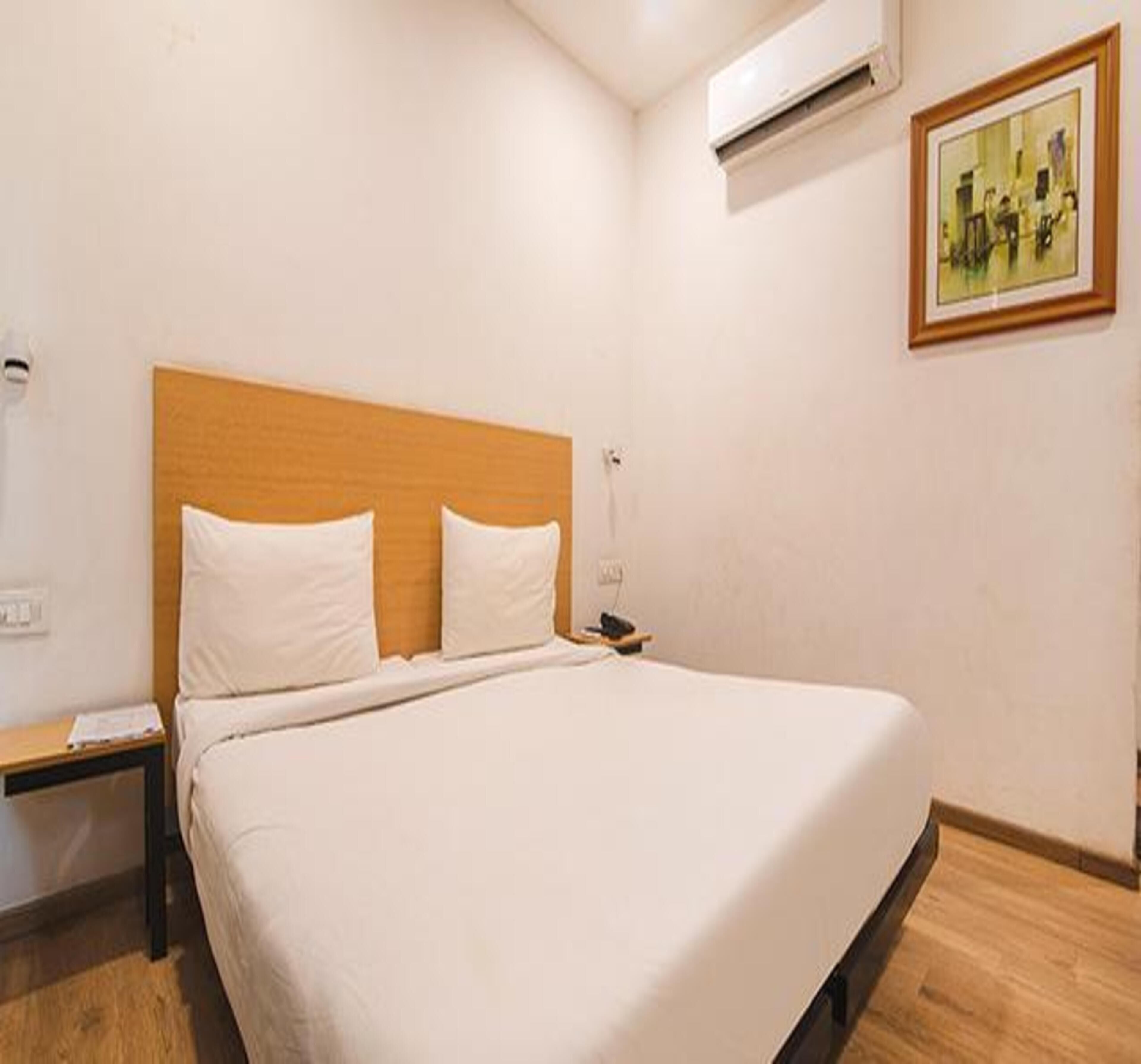 A hotel room with a single bed, a nightstand, an air conditioning unit, and a framed picture on the wall.