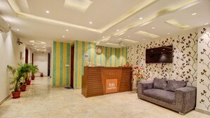 A reception area with a wooden desk, a sofa, potted plants, a clock, and a TV on the wall. - FabHotel Gross inn Suites (New Delhi)