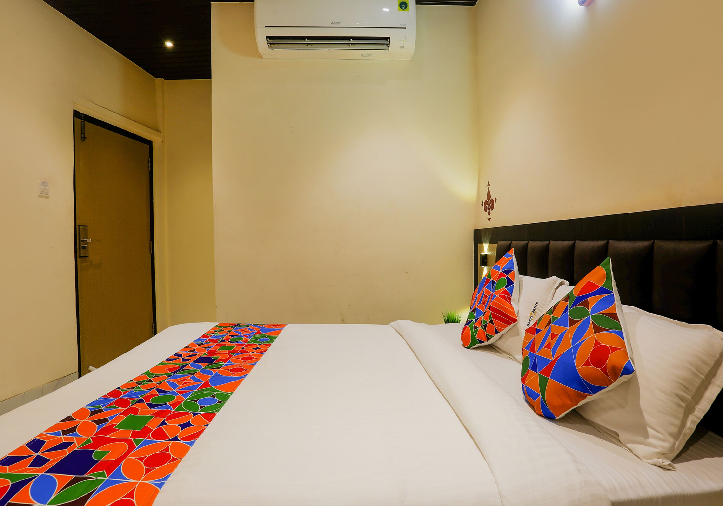A hotel room with a bed, a decorative pillow, a wall-mounted air conditioner, and a door.