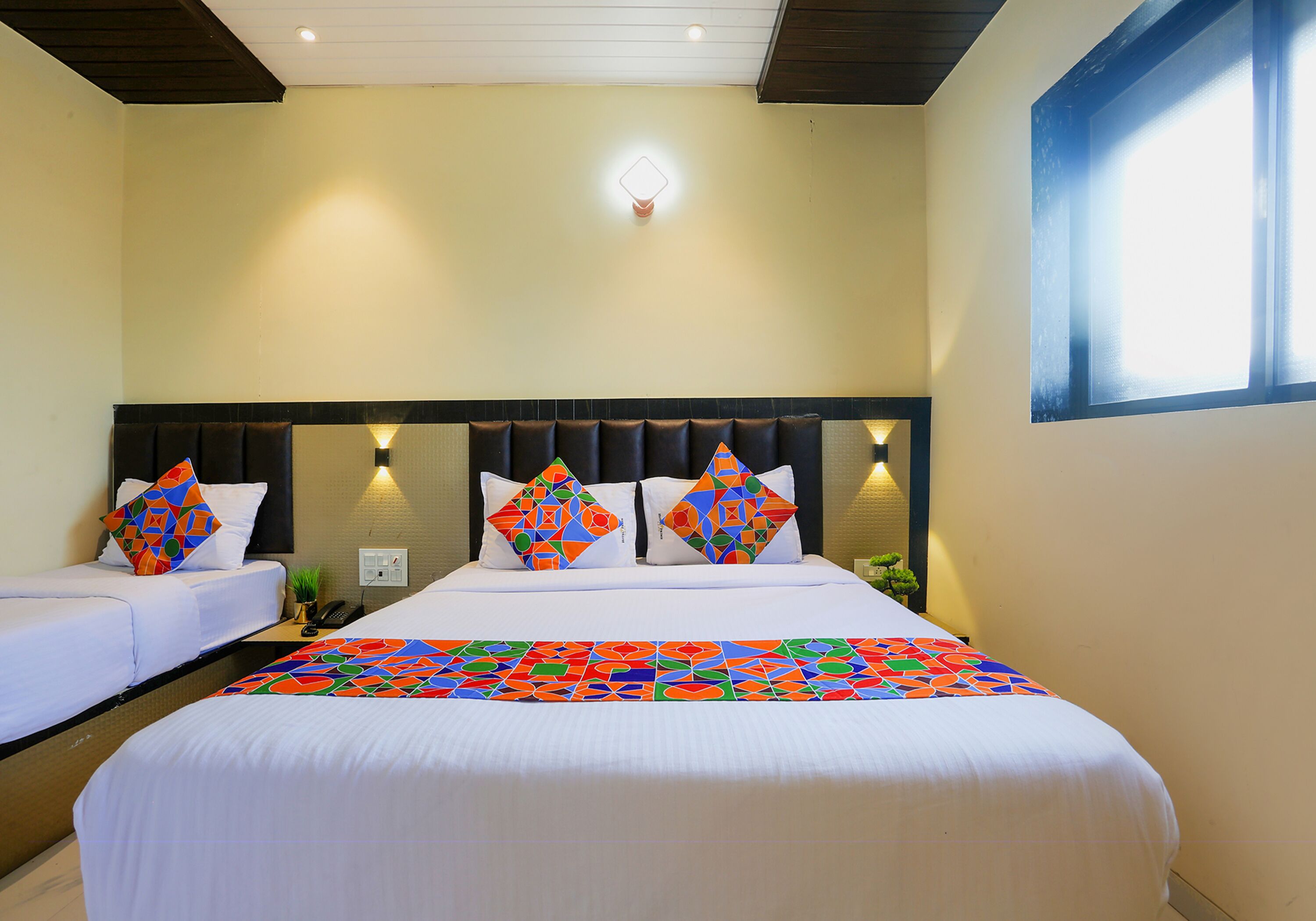 A hotel room with a large bed, two smaller beds, a headboard with lights, and a window with blinds.