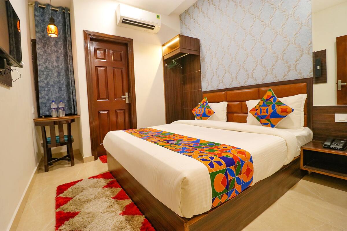 Quarto de hotel com cama, mesa de cabeceira com telefone, pequena mesa com garrafas de água, cabideiro e porta.