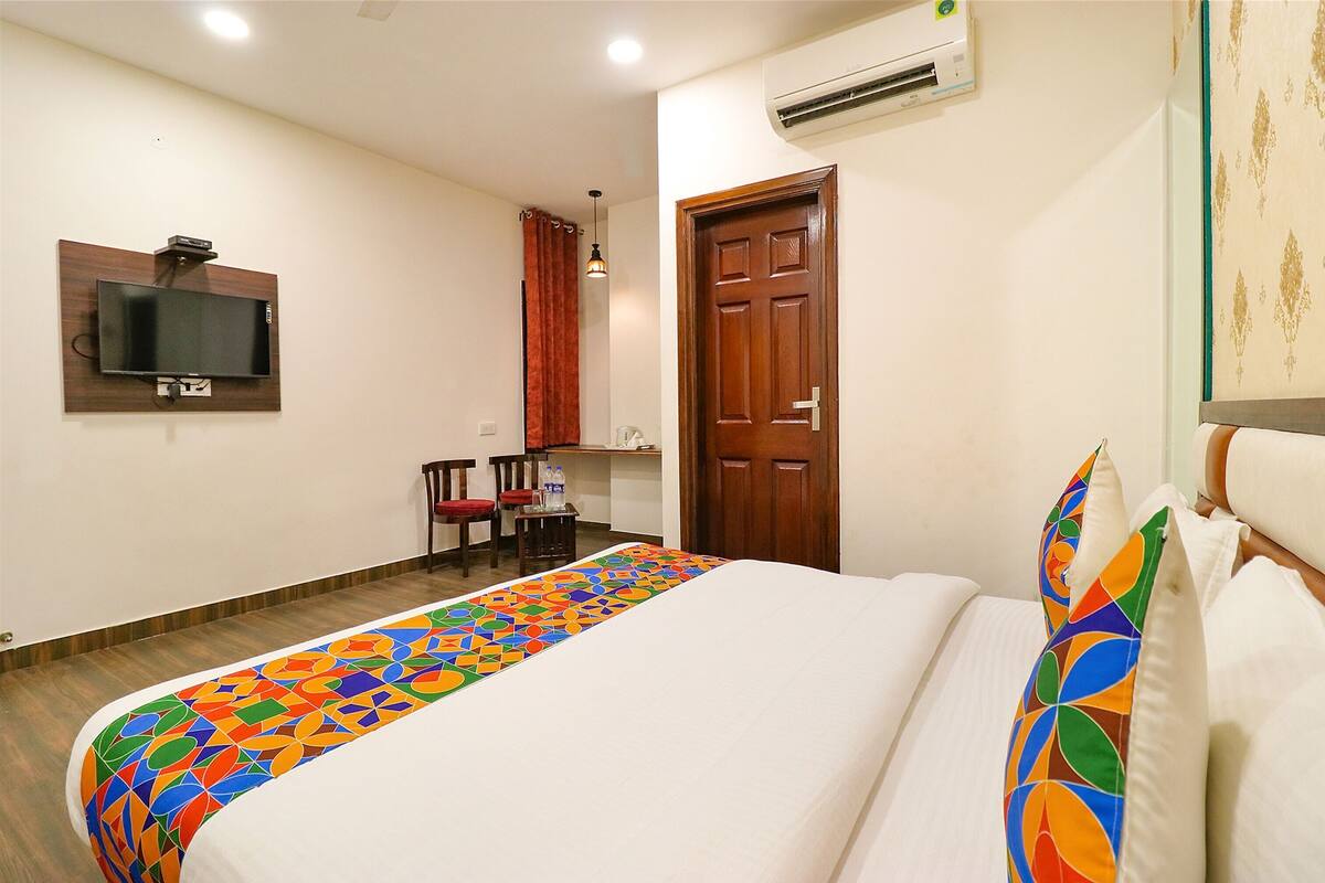 Quarto de hotel com cama, televisão na parede, porta de madeira e pequena mesa com cadeiras.