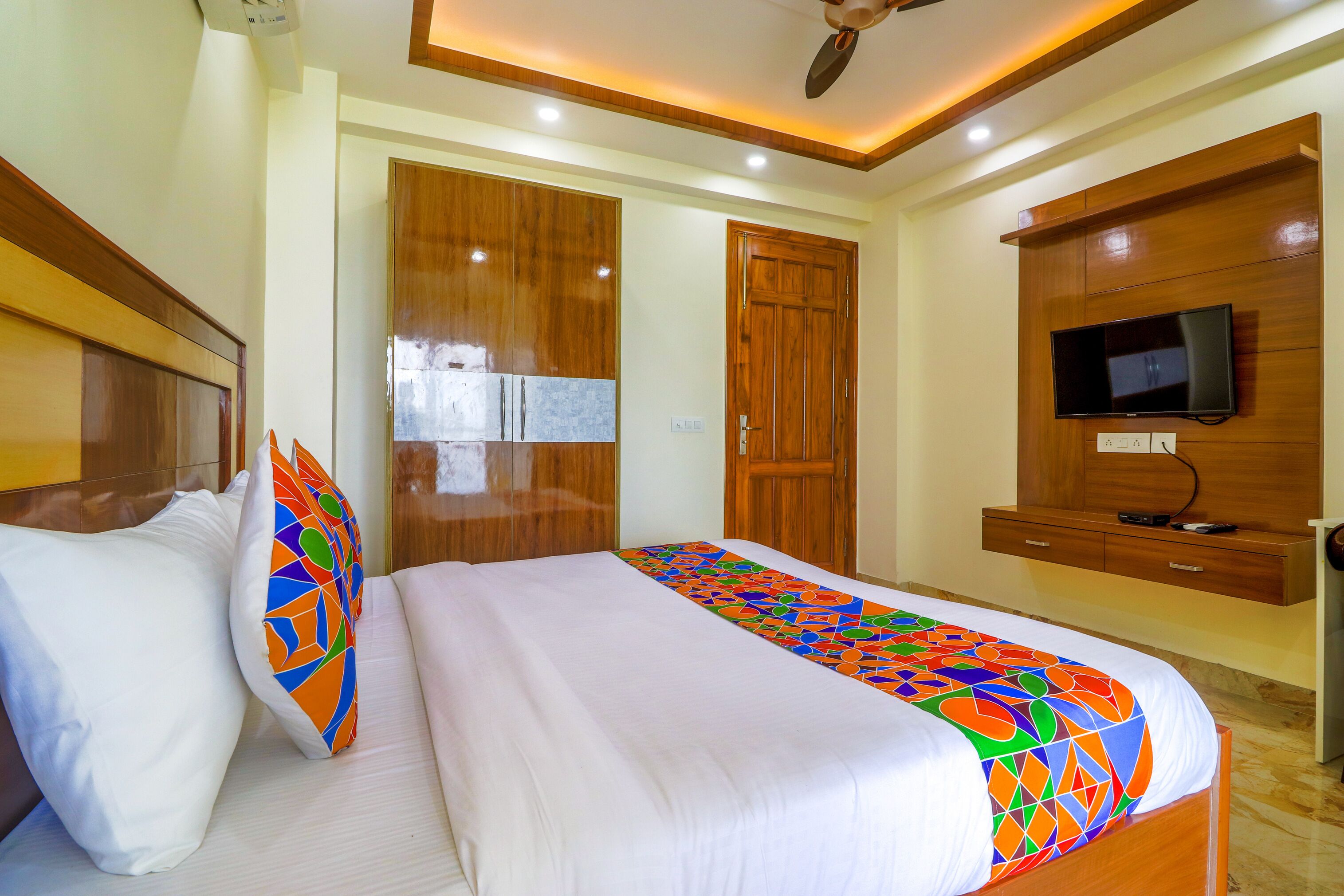 A hotel room with a large bed, wooden wardrobe, a television mounted on the wall, and a ceiling fan.