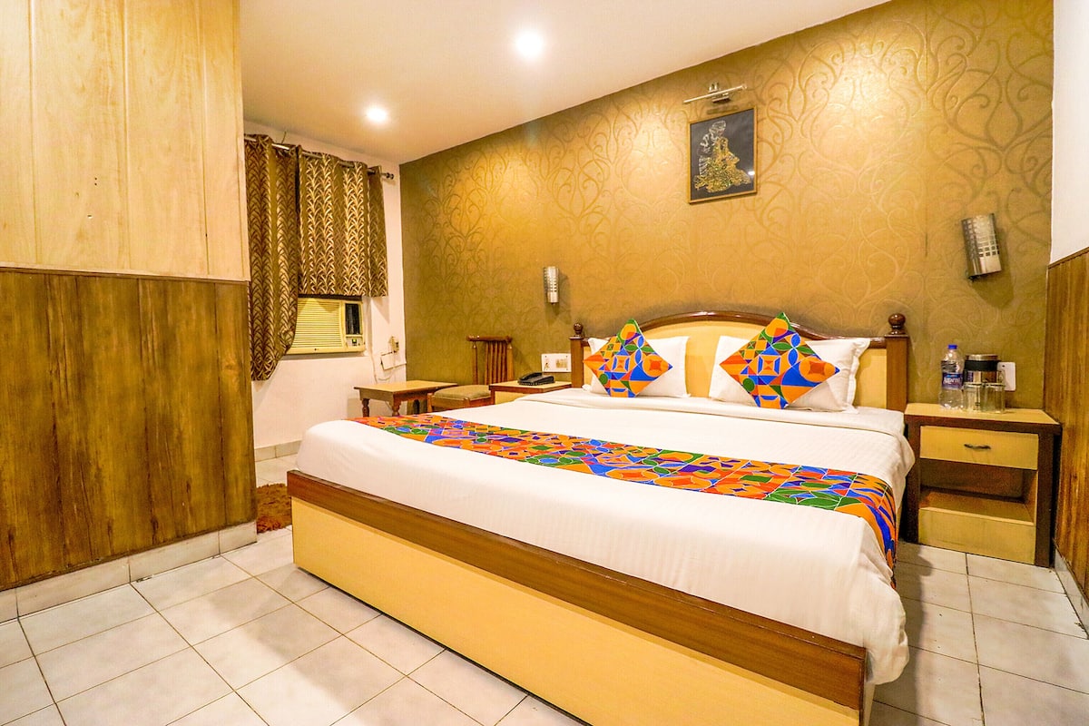 A hotel room with a large bed, a wooden wardrobe, a bedside table, a nightstand, and a wall-mounted lamp.