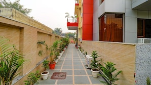 A corridor lined with potted plants, leading to a building with a red and grey facade. - FabHotel Elite Peak (Noida)