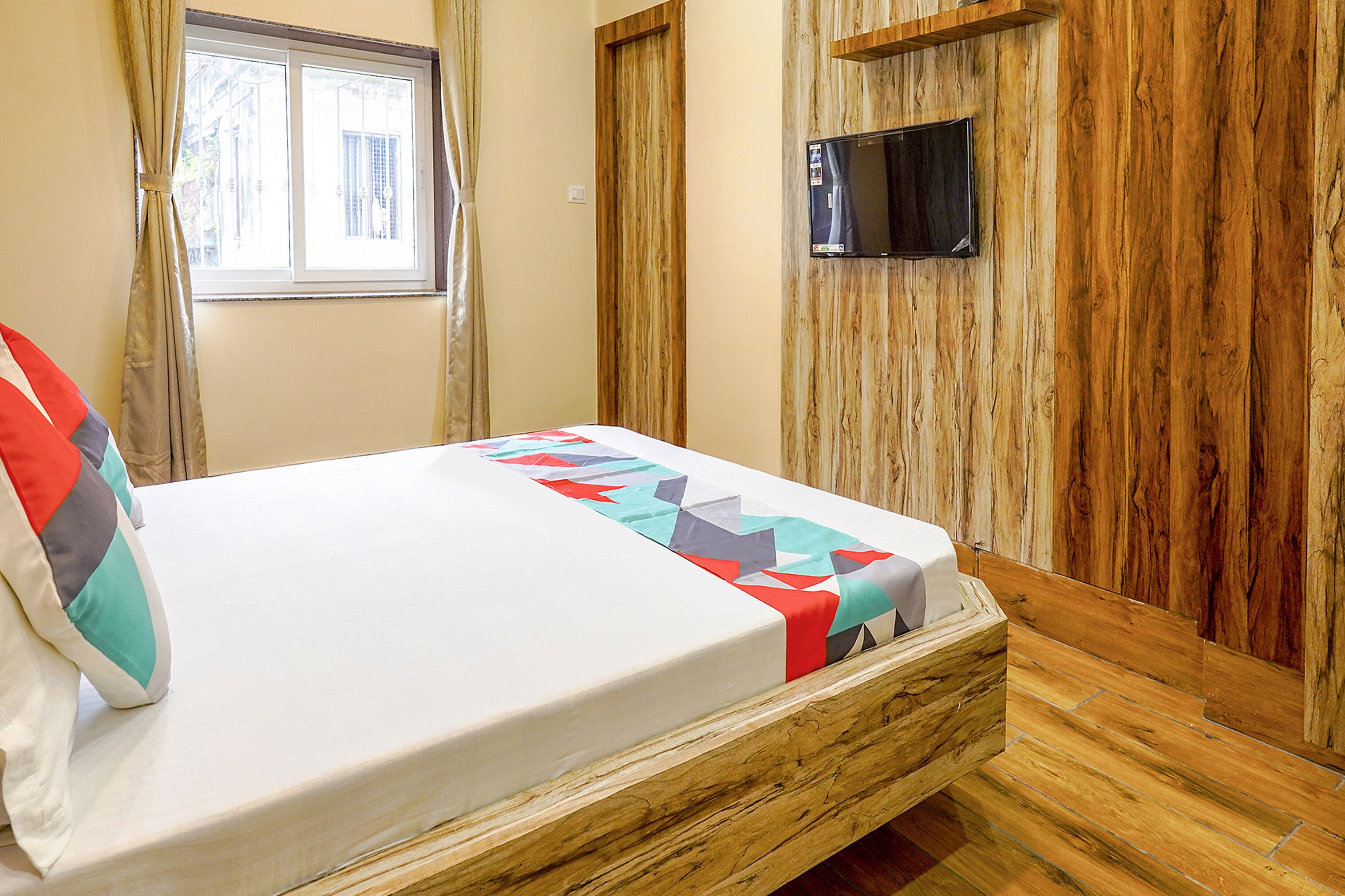 A bed with a white mattress and colorful quilt, a wooden headboard, a TV mounted on the wall, and a window with curtains.