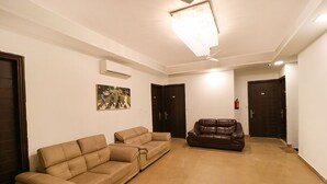 A spacious living area with two beige sofas and a brown leather sofa, a ceiling light fixture, and a wall-mounted air conditioner. - Fabhotel The Royal Oak (Gurugram)