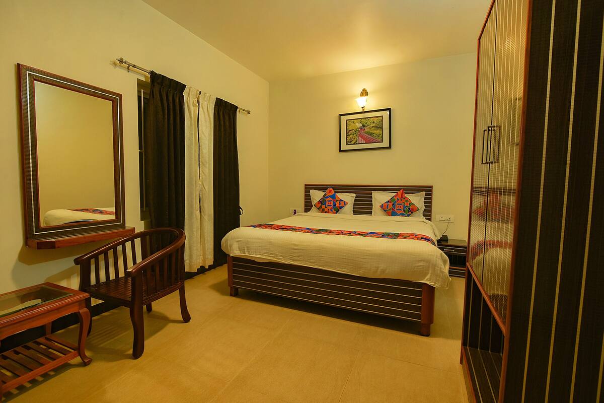 Um quarto com cama, cadeira de madeira, mesa de cabeceira, espelho e guarda-roupa.