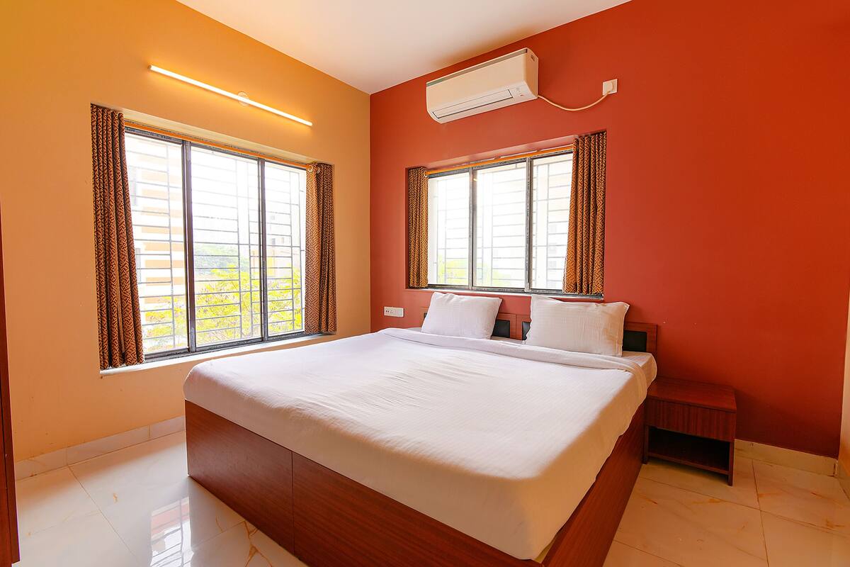A hotel room with a large bed, two windows with curtains, an air conditioning unit, and a small bedside table.