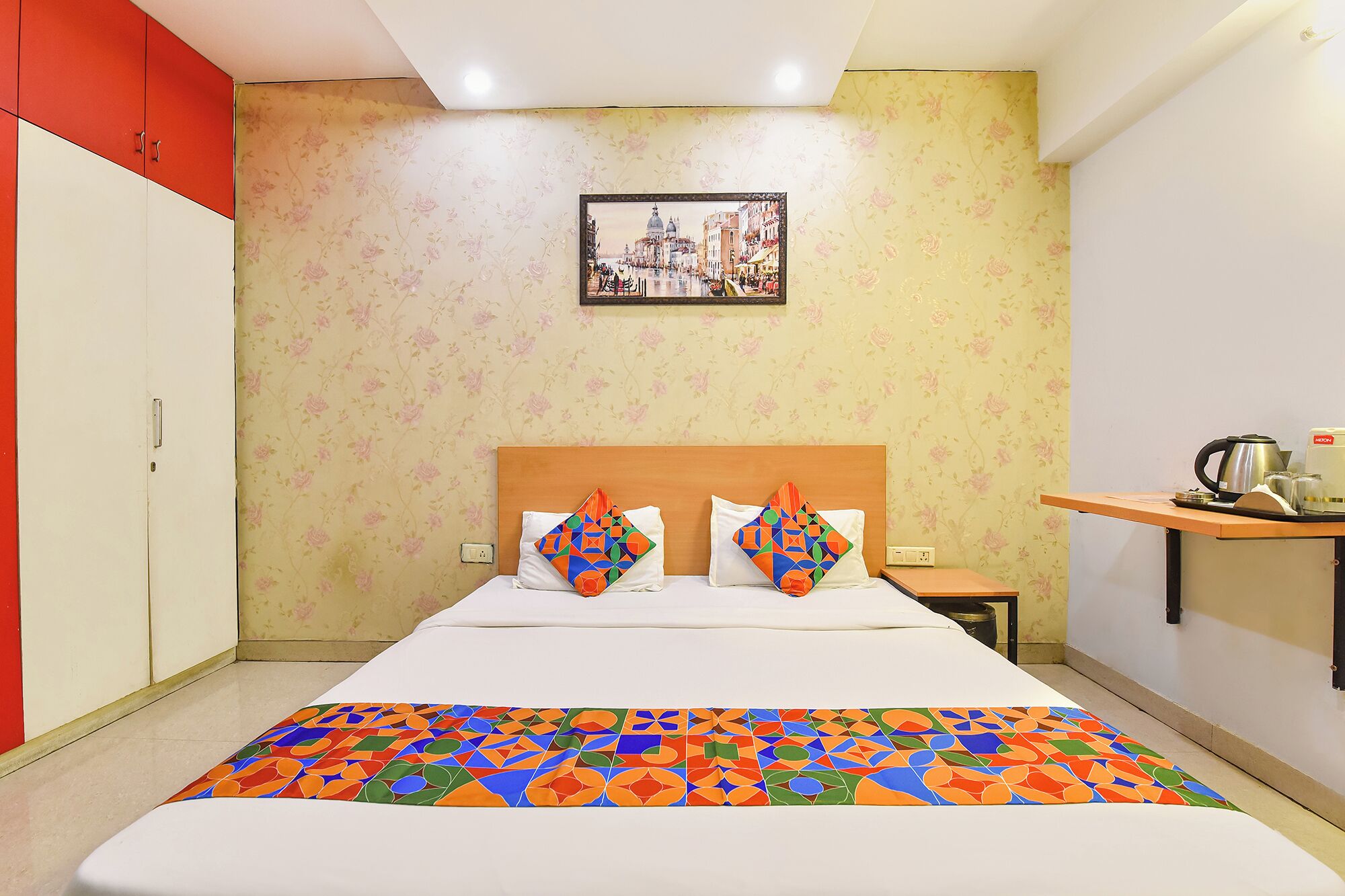 A hotel room with a bed, a nightstand, a red wardrobe, a floral wallpaper, and a cityscape painting.