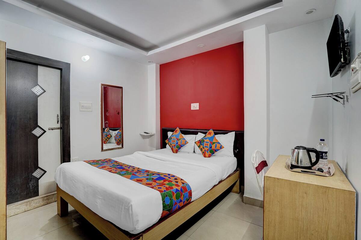 Habitación de hotel con una cama, una mesita de noche de madera, una pared de acento roja, un espejo y un televisor montado en la pared.