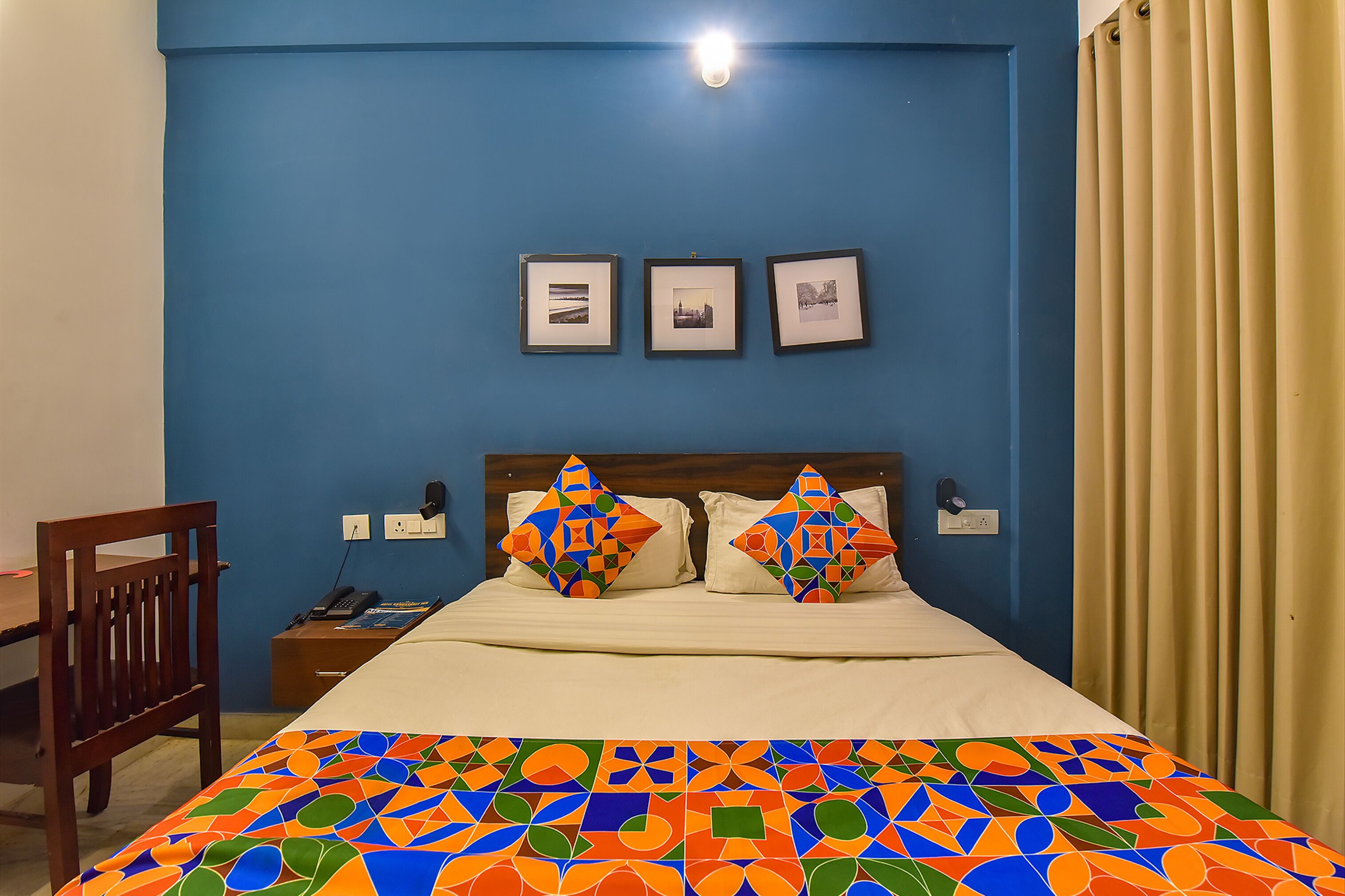 A bedroom with a blue wall, a bed with a colorful geometric-patterned bedspread, two decorative pillows, and three framed pictures on the wall.