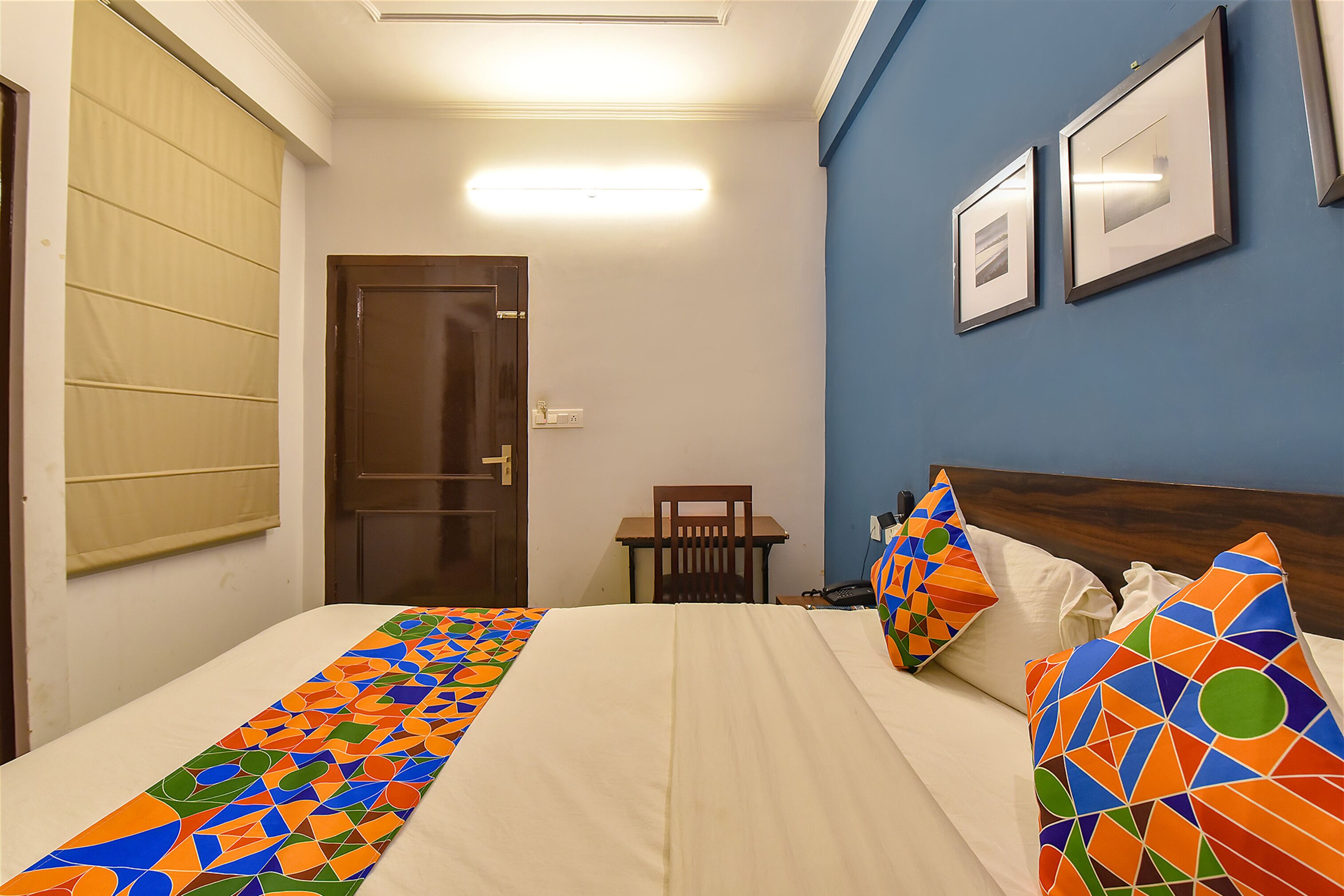 A hotel room with a bed, a chair, a table, and two framed pictures on the wall.