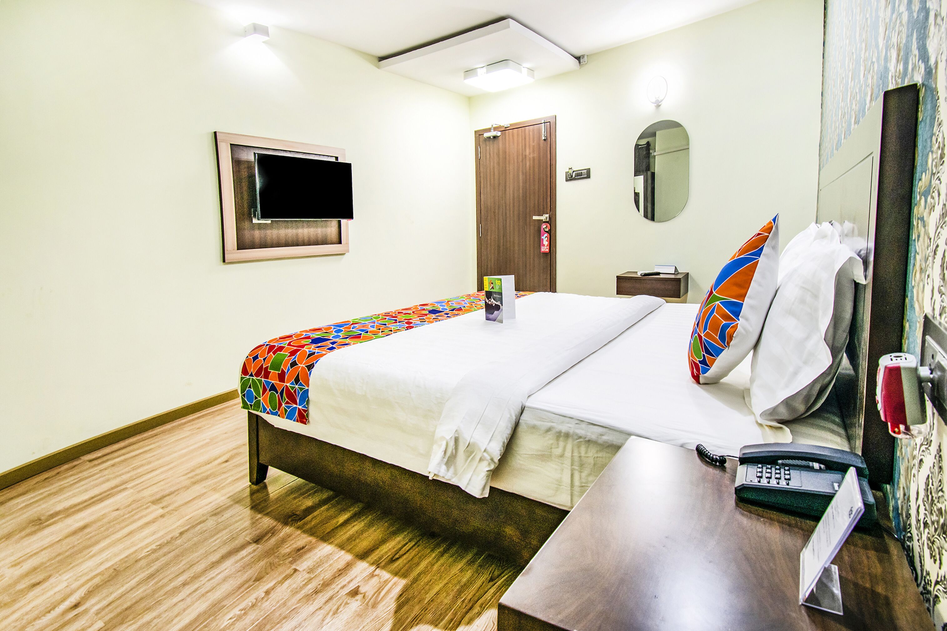 A hotel room with a bed, a bedside table with a phone, a TV mounted on the wall, a door, and a mirror.