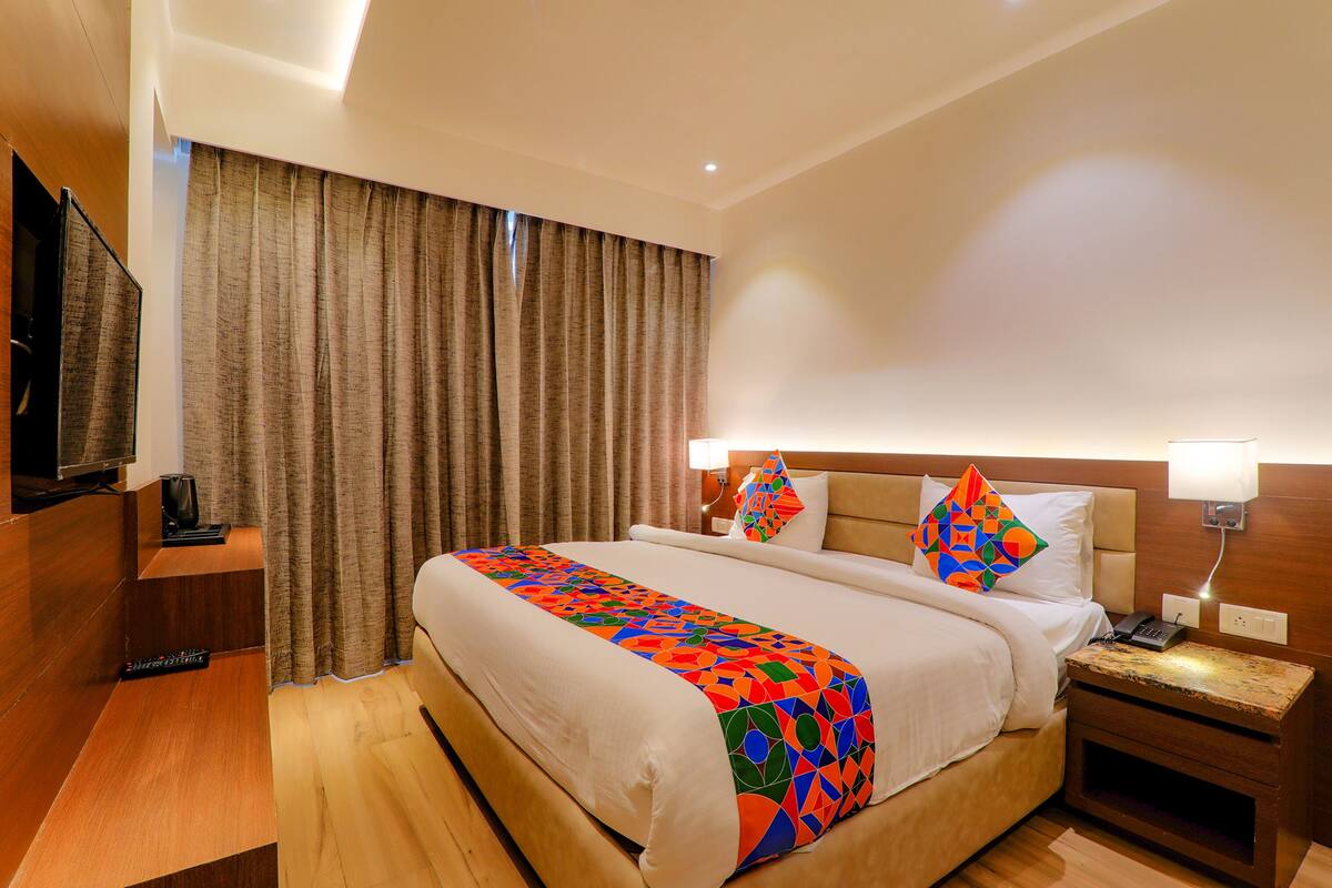 Habitación de hotel con cama, televisión, mesita de noche, lámpara y ventana con cortinas.