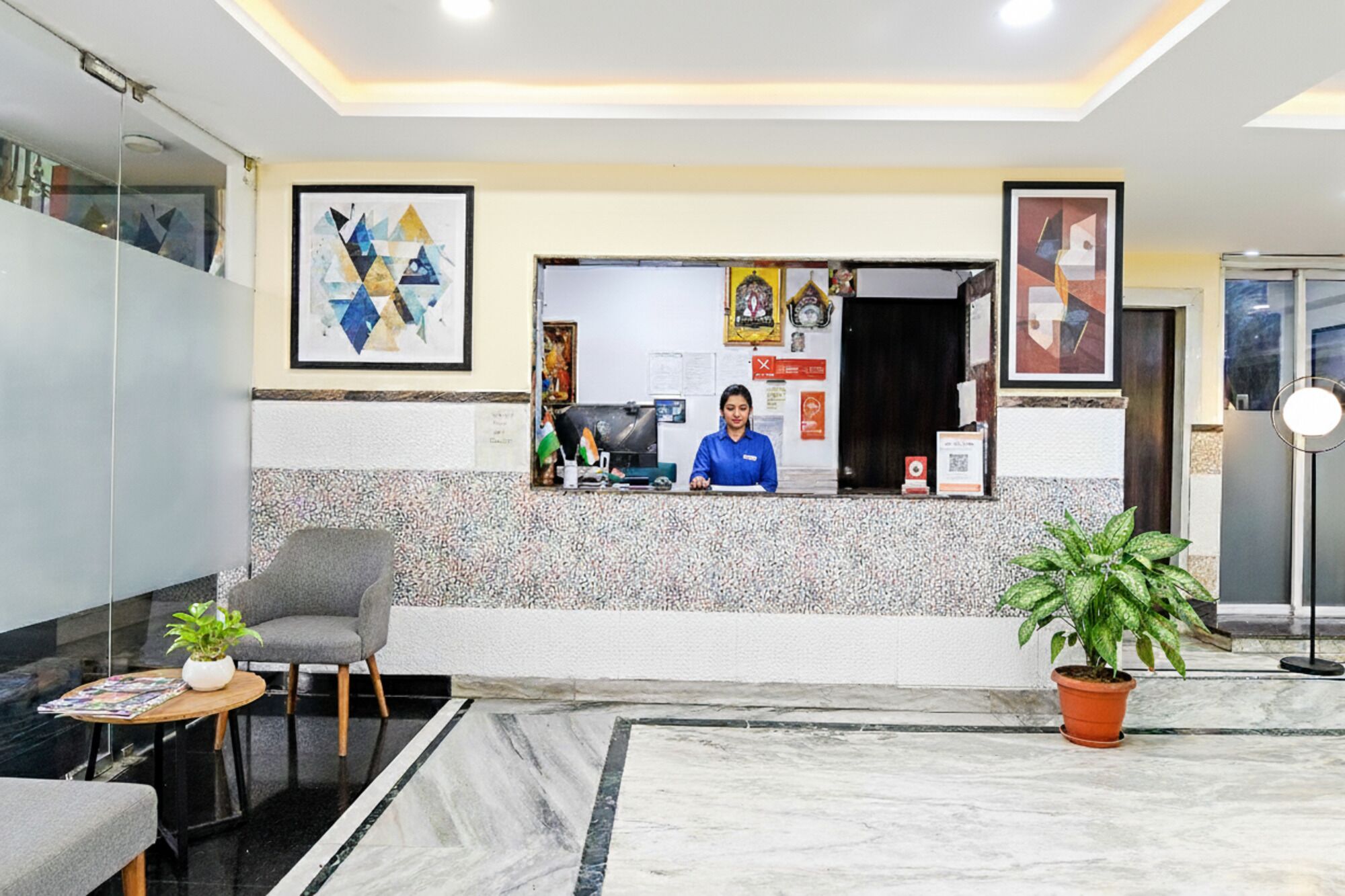 A reception area with a receptionist, a marble floor, a chair, a small table with a plant, and framed artwork on the walls.