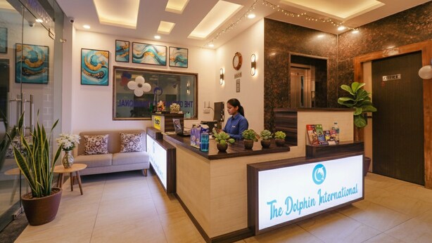 A reception desk with a sign reading "The Dolphin International". There is a receptionist behind the desk, a sofa with cushions, and a potted plant to the left. - FabHotel Dolphin International (New Delhi)