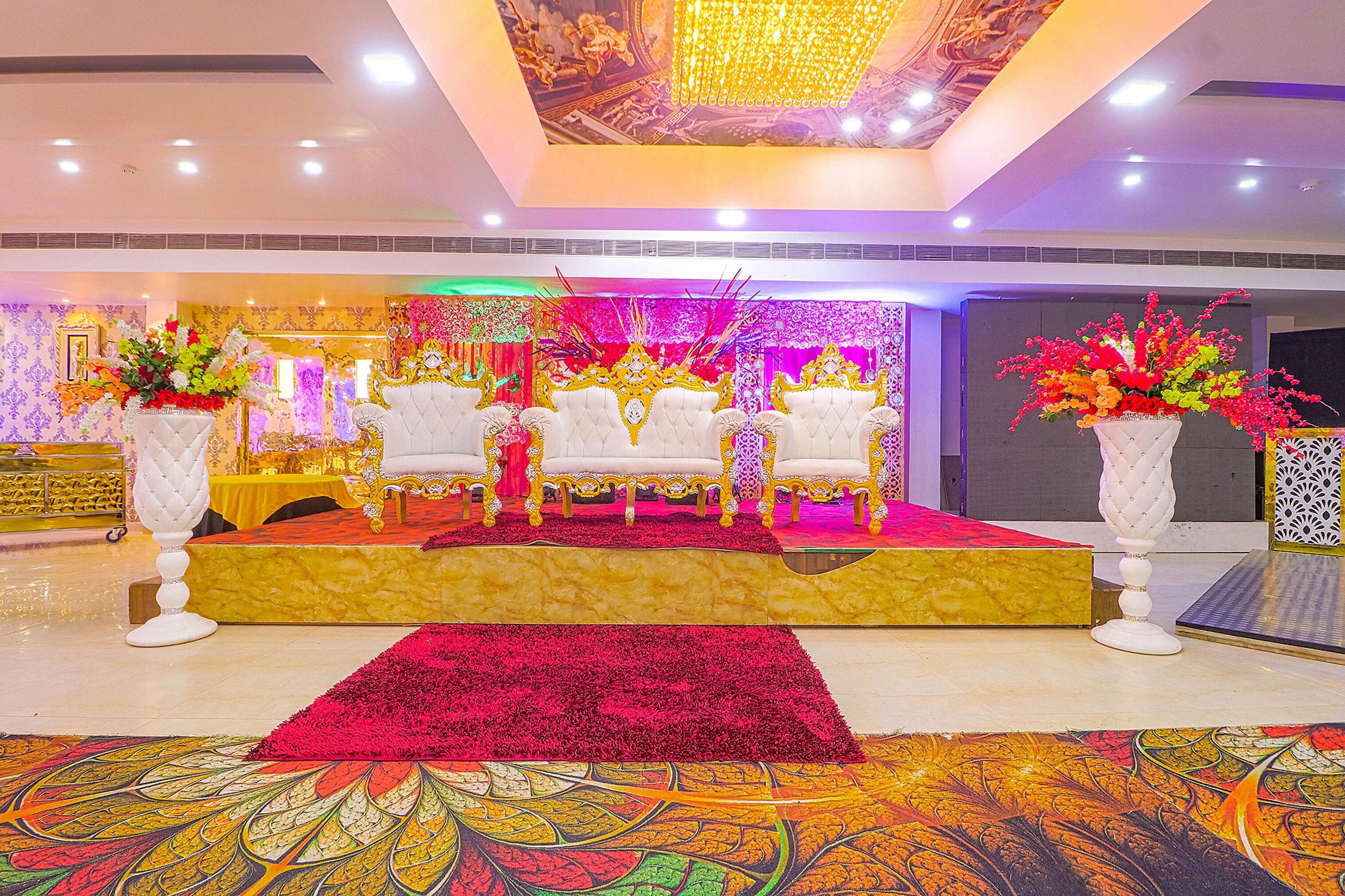 Un escenario con sillas blancas y una alfombra roja, rodeado de arreglos florales y un suelo con diseño.