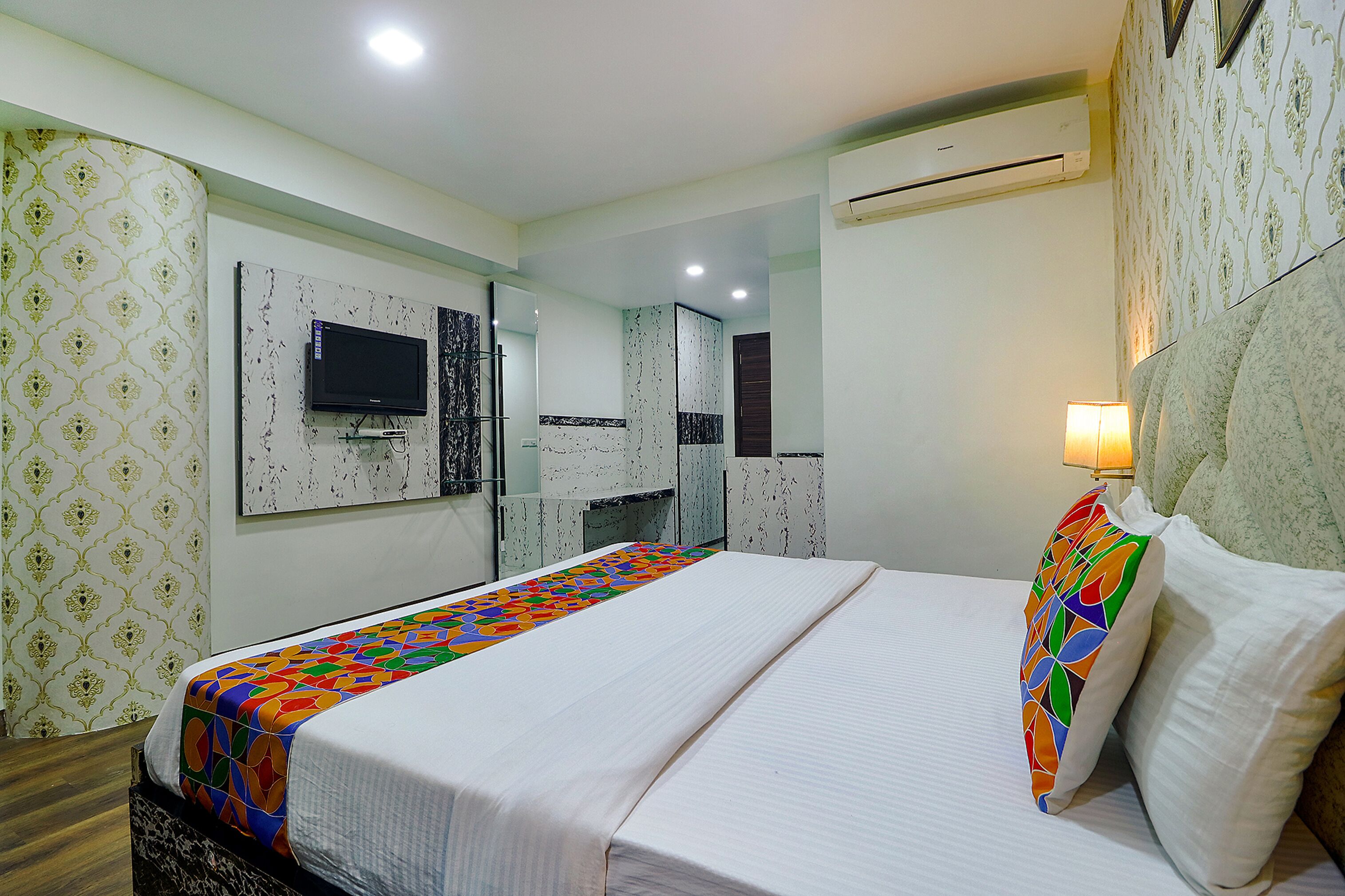 A hotel room with a bed, a TV, a mirror, and a wall-mounted air conditioner.