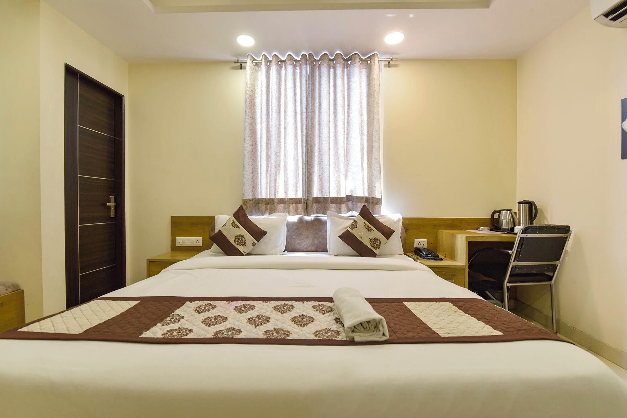 A hotel room with a bed, a desk, a chair, a window with curtains, and a coffee maker.