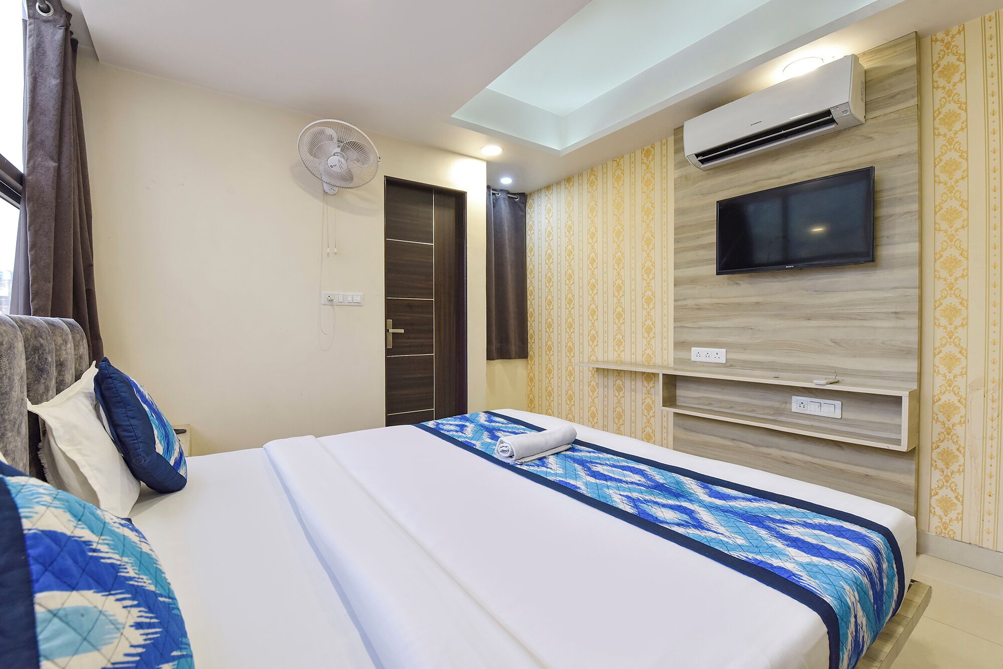 A hotel room with a bed, a TV mounted on the wall, an air conditioning unit, a ceiling fan, and a door with a curtain.