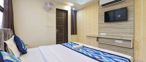 A hotel room with a bed, a TV mounted on the wall, an air conditioning unit, a ceiling fan, and a door with a curtain.