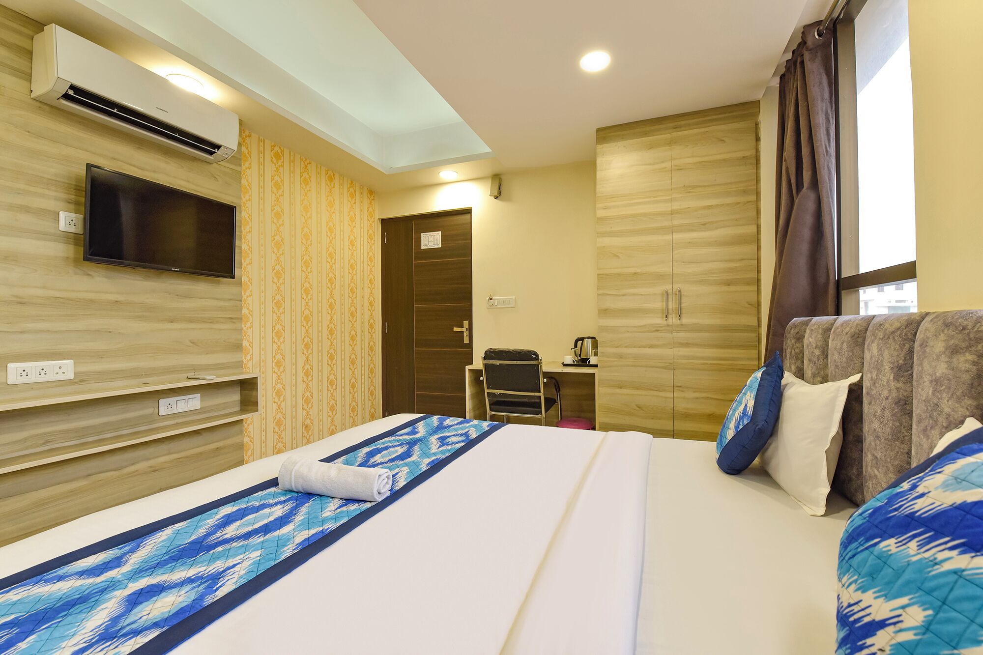 A hotel room with a bed, a TV mounted on the wall, a desk with a chair, and a wardrobe.