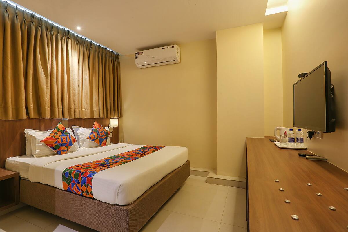 A hotel room with a bed, a desk, a flat-screen TV, an air conditioner, and a window with curtains.
