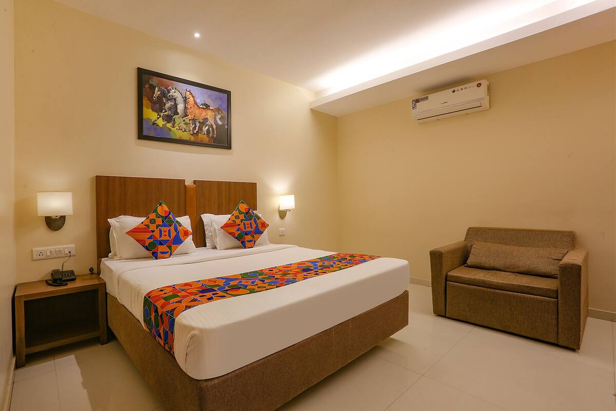 A hotel room with a bed, a chair, a nightstand, a wall-mounted air conditioner, and a painting on the wall.