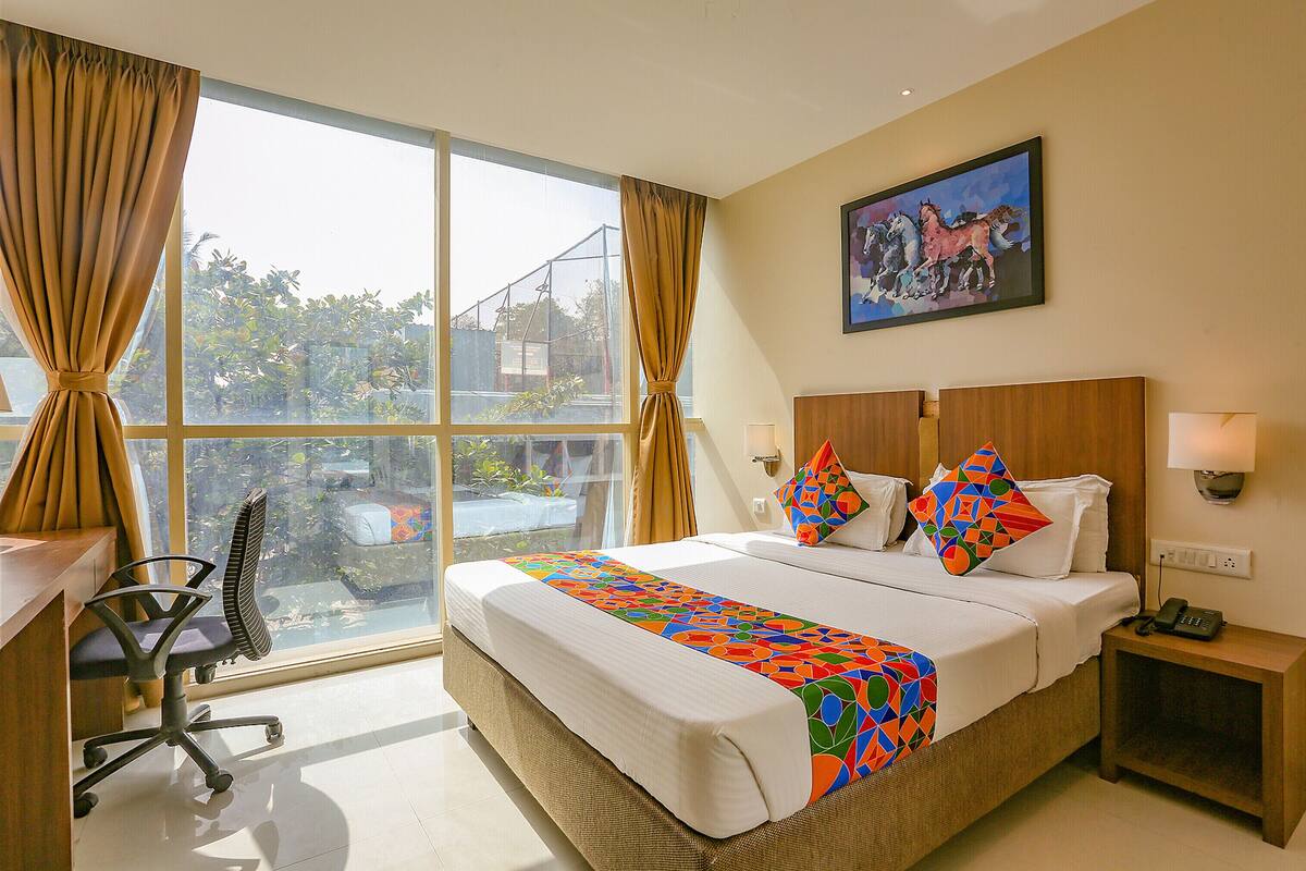 A hotel room with a large window, a bed with colorful bedding, a desk with a chair, and a painting on the wall.